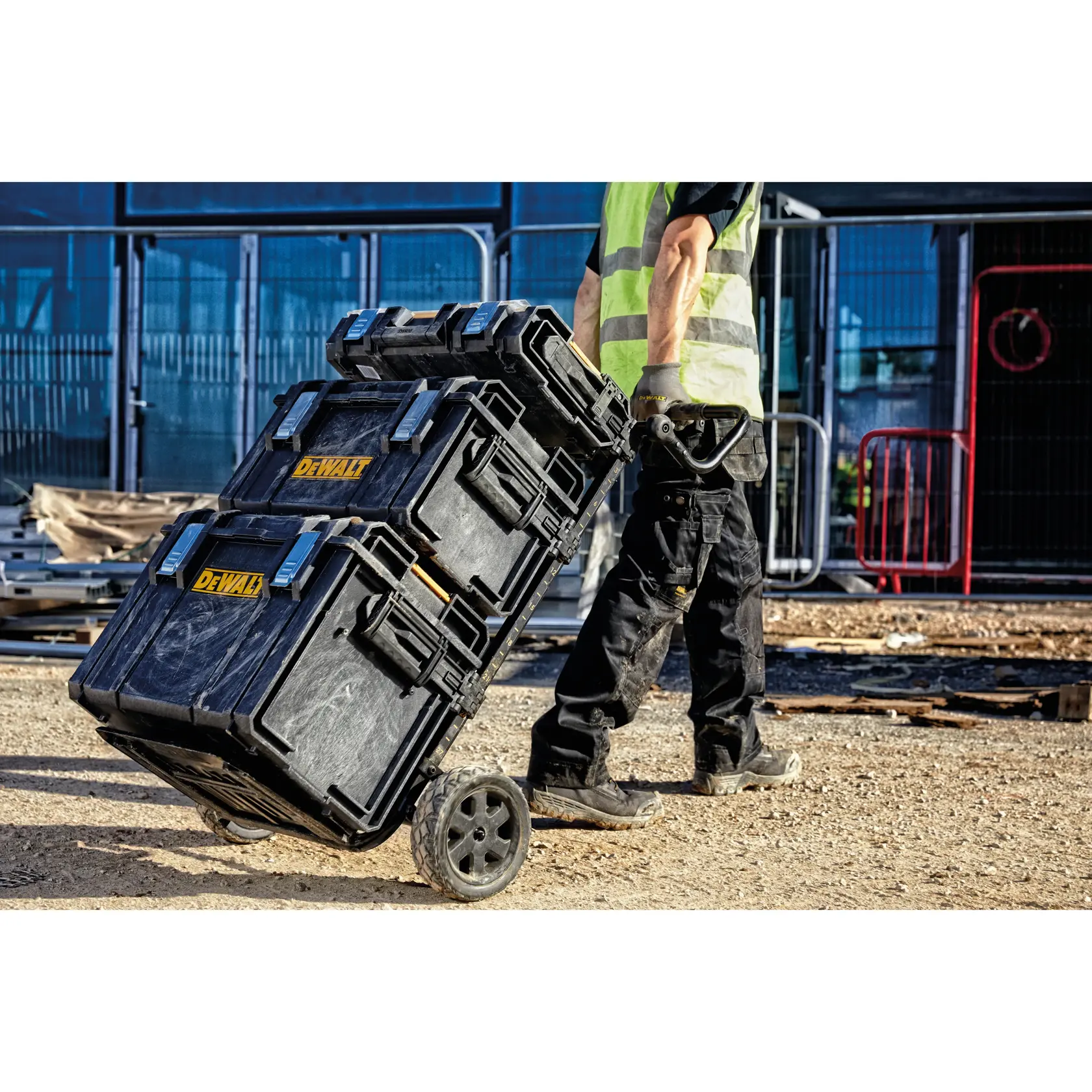 Eine Person in Arbeitskleidung und Warnweste zieht drei schwarze DEWALT-Werkzeugkisten auf einem Rollwagen über eine Baustelle.