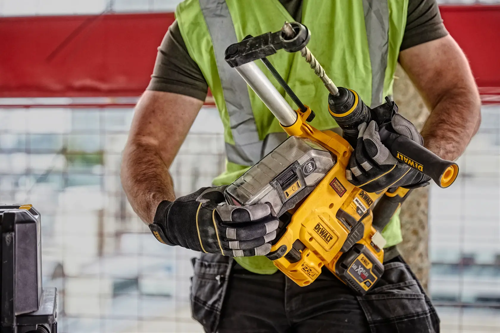 Eine Person mit grüner Sicherheitsweste und Handschuhen hält einen DEWALT D25304DH Akku-Bohrhammer mit angeschlossenem Staubabsaugsystem auf einer Baustelle.