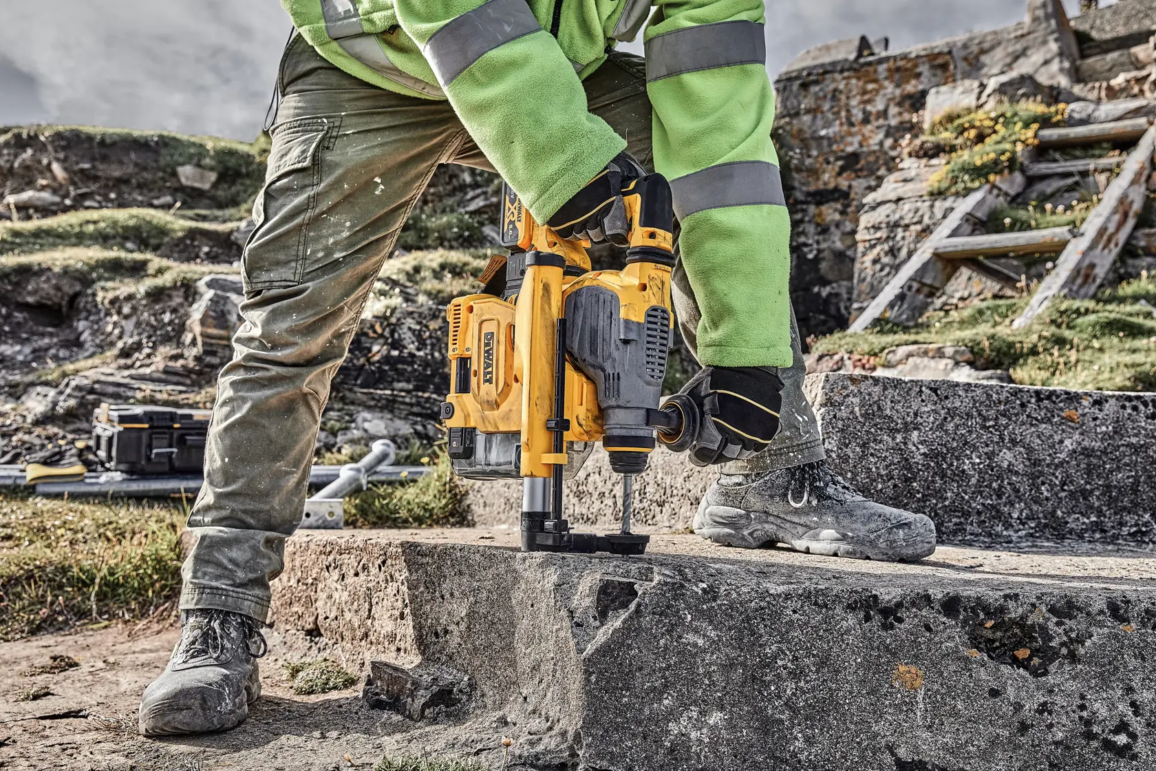 Person nutzt DEWALT-Bohrer auf Beton im Freien, mit Handschuhen, Schuhen und Schutzkleidung.
