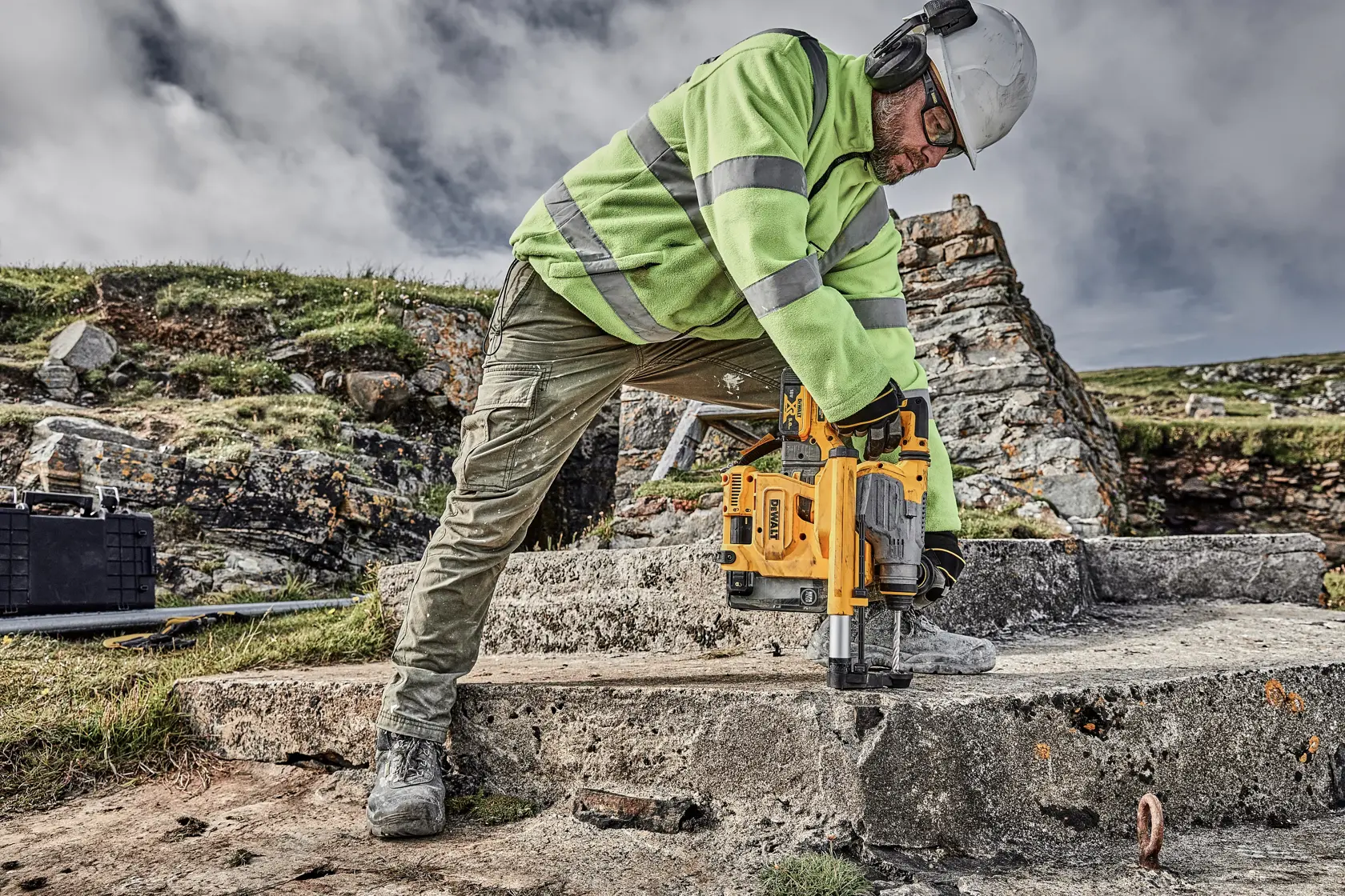 Arbeiter bohrt draußen mit gelbem DEWALT-Werkzeug in Beton, trägt Schutzkleidung und Warnjacke.