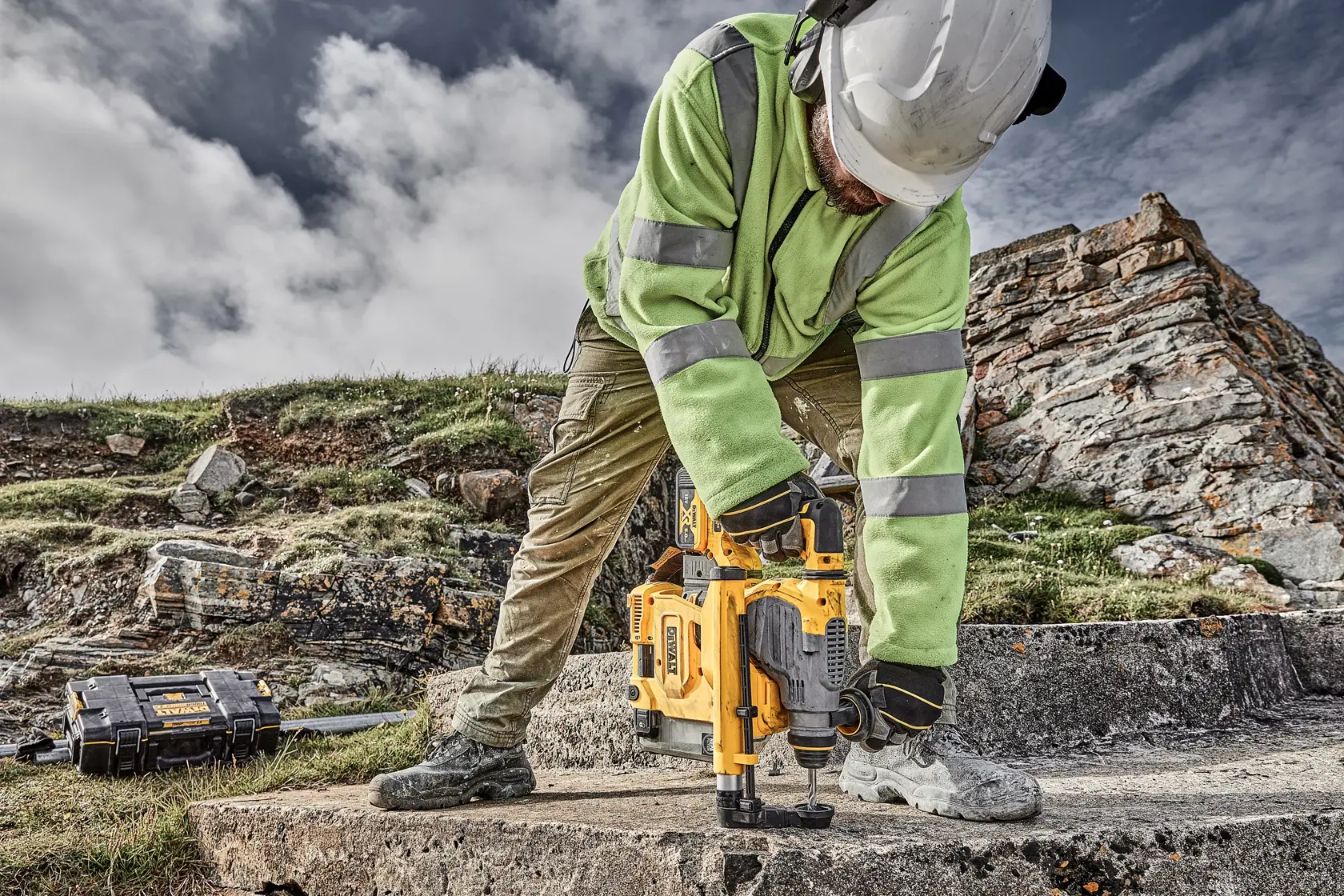 Arbeiter nutzt DEWALT Werkzeug auf Beton im Freien, mit Schutzkleidung und Helm, Werkzeugkiste in Nähe.