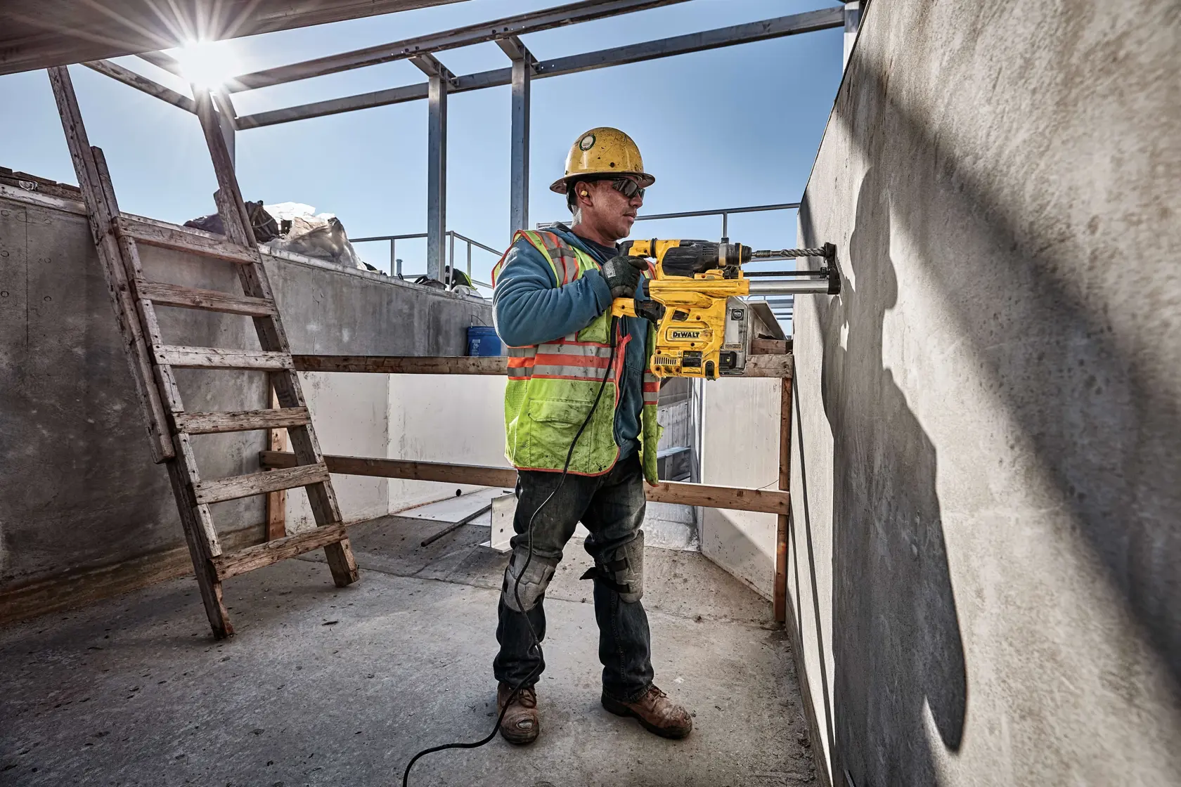 A construction worker wearing safety gear operates a DEWALT D25333K rotary hammer drill against a concrete wall at an outdoor job site.