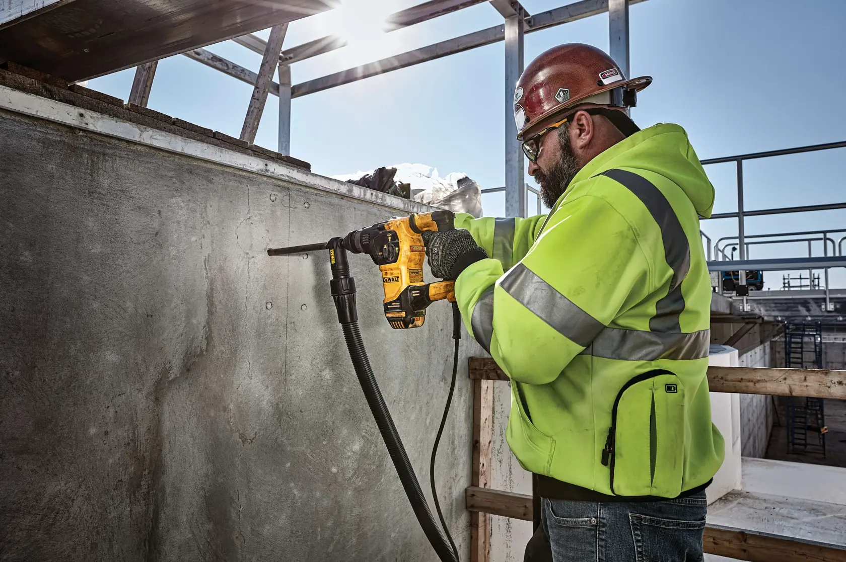 A construction worker wearing a safety vest and helmet is using a DEWALT D25333K rotary hammer drill to drill into a concrete wall at a construction site. The worker's face is blurred for privacy.