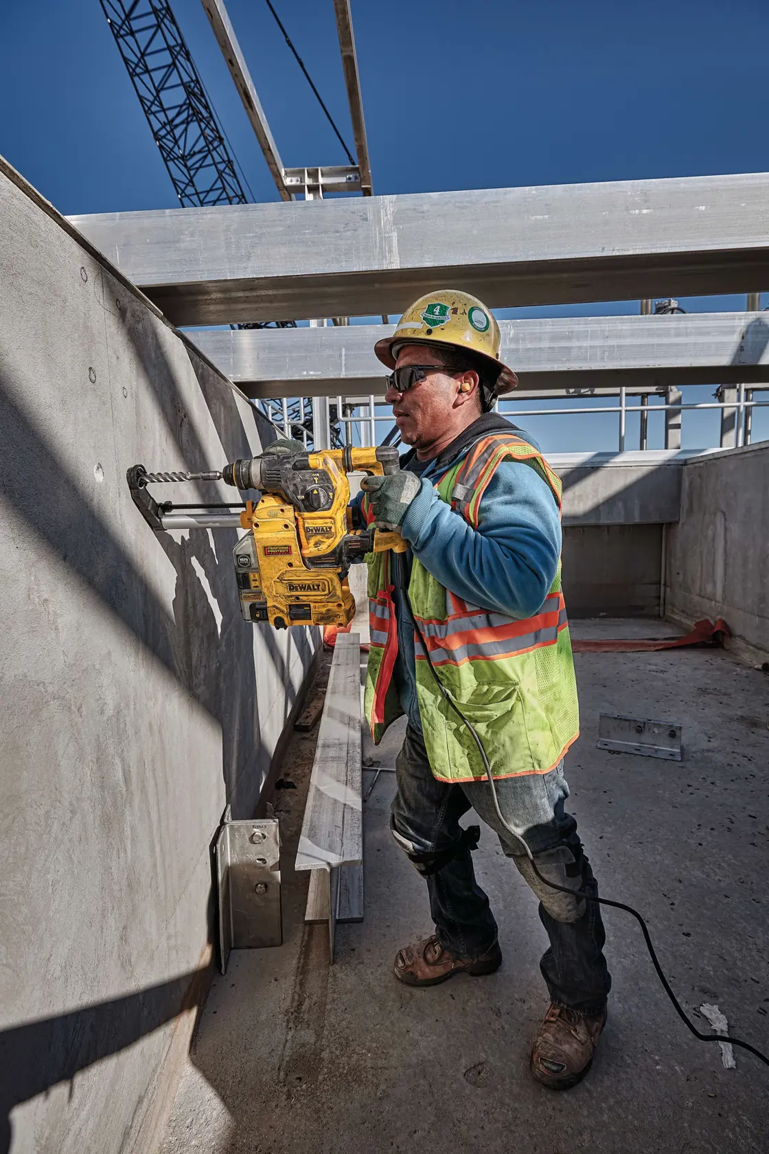 Un ouvrier du bâtiment portant un casque de sécurité et un gilet haute visibilité utilise une perceuse à percussion rotative jaune DEWALT, modèle D25333K, pour percer un trou dans un mur en béton sur un chantier en plein air. L’ouvrier se tient près d’une échelle en bois sous une structure partiellement construite, avec le soleil éclatant.