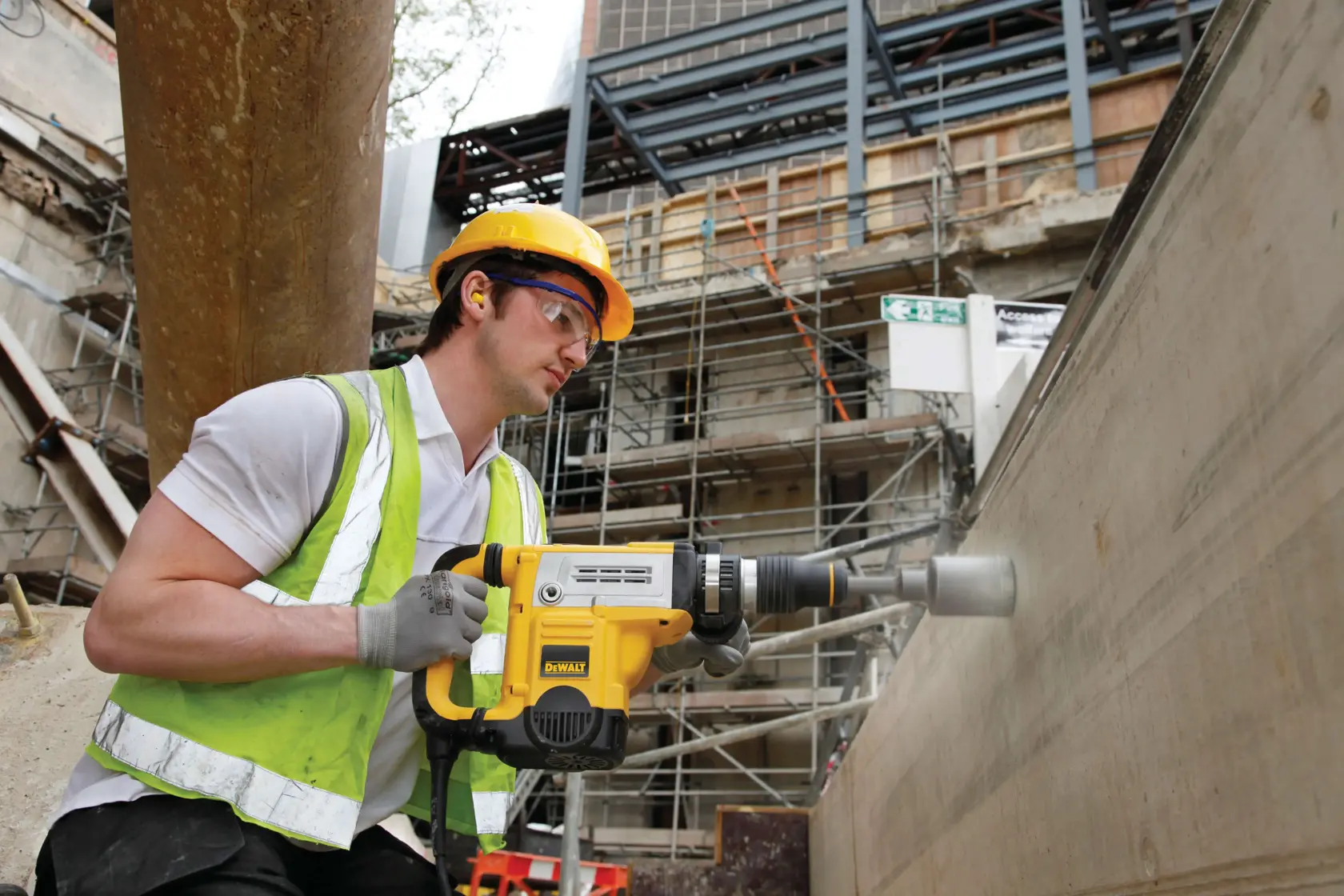 Ein Bauarbeiter mit gelbem Schutzhelm und reflektierender Weste verwendet einen DEWALT D25601K Bohrhammer an einer Betonwand auf einer Baustelle mit Gerüsten und Baumaterialien im Hintergrund.