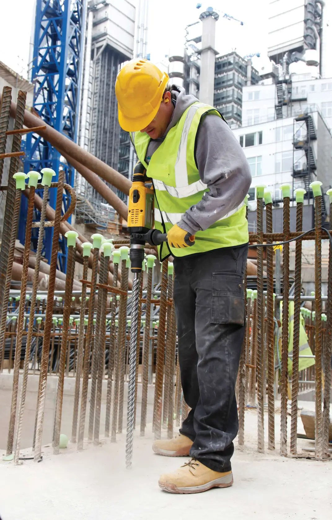Eine Person mit gelbem Schutzhelm, Sicherheitsweste, Handschuhen und Arbeitsstiefeln verwendet einen DEWALT Drehhammer-Bohrmaschine (Modell D25601K), um Beton auf einer Baustelle mit Metallbewehrung zu bohren. Im Hintergrund sind moderne Gebäude und Baukräne zu sehen.