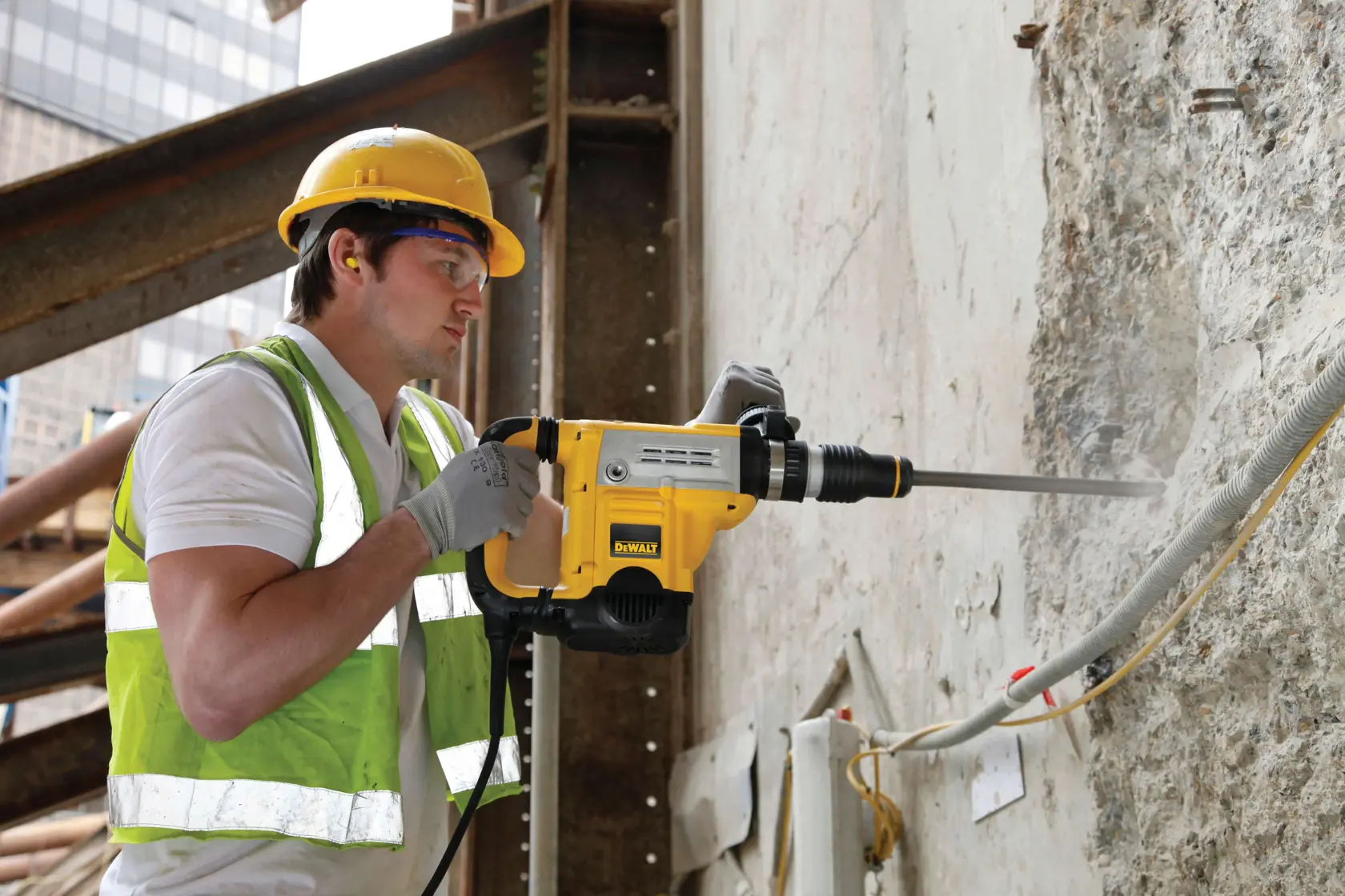 Ein Bauarbeiter mit gelbem Schutzhelm, Handschuhen und reflektierender Weste verwendet einen DEWALT Elektrodrehhammer (D25601K), um eine Betonwand auf einer Baustelle zu bearbeiten.