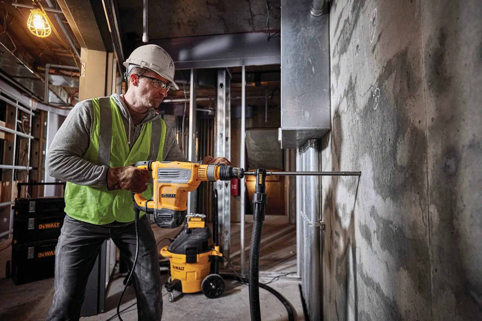 Ein Arbeiter mit Sicherheitshelm und Weste bedient einen DEWALT Drehhammer-Bohrer (SKU: D25614K), um eine Betonwand auf einer Baustelle zu bohren. DEWALT Geräte und Werkzeuge sind im Hintergrund sichtbar.