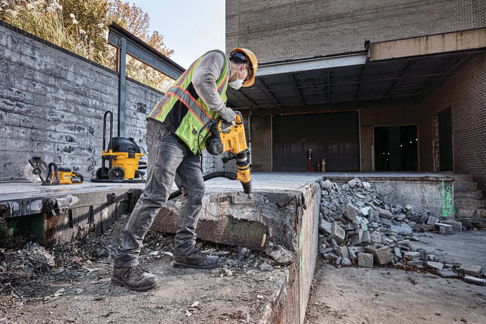 Ein Bauarbeiter mit Schutzausrüstung und reflektierender Weste benutzt einen DEWALT Abbruchhammer, um Beton auf einer Baustelle im Freien zu zerkleinern. Weitere DEWALT Werkzeuge und Ausrüstung sind im Hintergrund sichtbar.