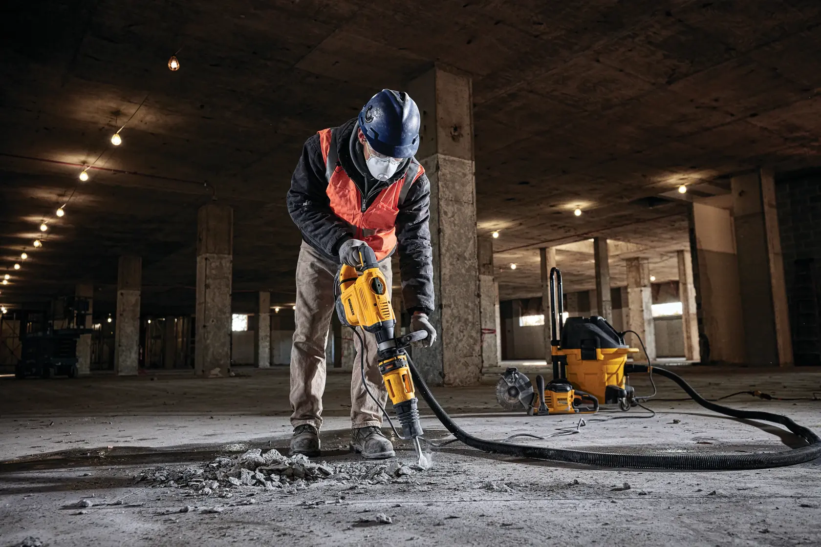 Ein Arbeiter mit Schutzkleidung bedient einen DEWALT Abbruchhammer (SKU: D25832K), um Beton auf dem Boden eines großen, unfertigen Gebäudes aufzubrechen. Ein gelber Industriestaubsauger ist über einen Schlauch mit dem Werkzeug verbunden und im Hintergrund zu sehen.