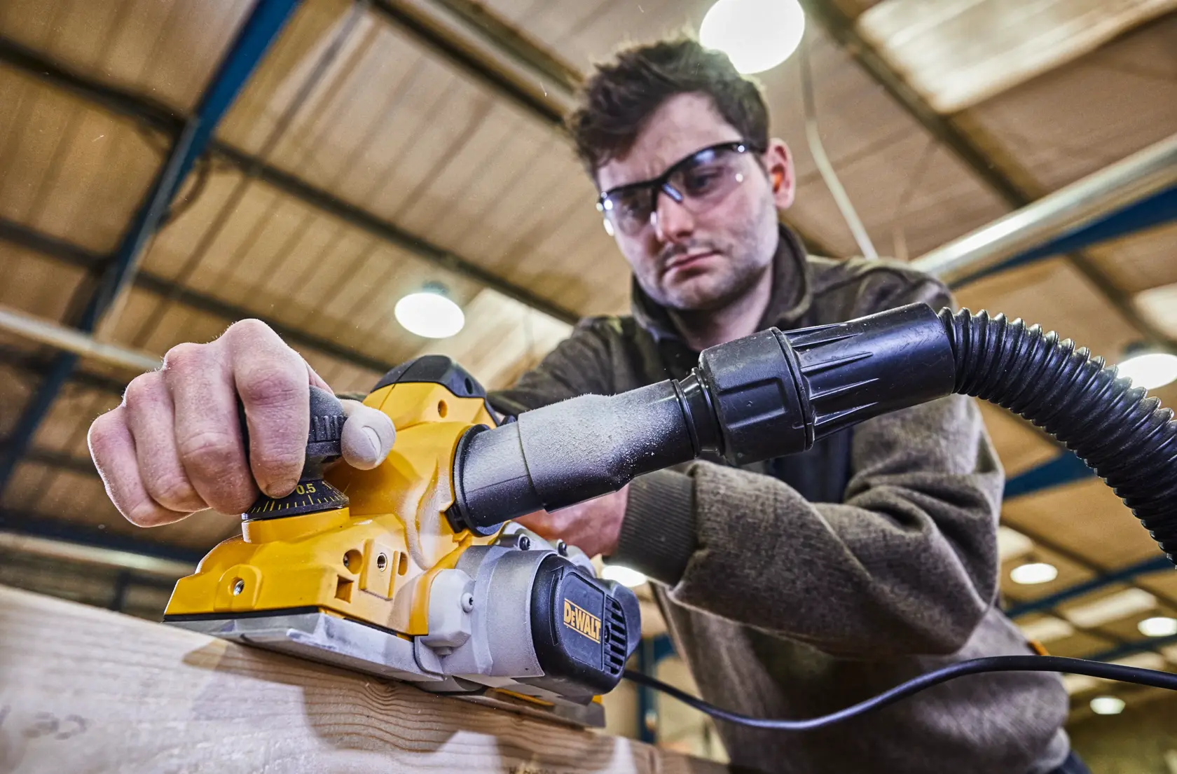 Eine Person verwendet einen DEWALT Elektrohobel (Modell D26500K) auf einem Holzstück in einer Werkstatt, mit angeschlossenem Staubabsaugschlauch. Das Gesicht der Person ist verwischt.