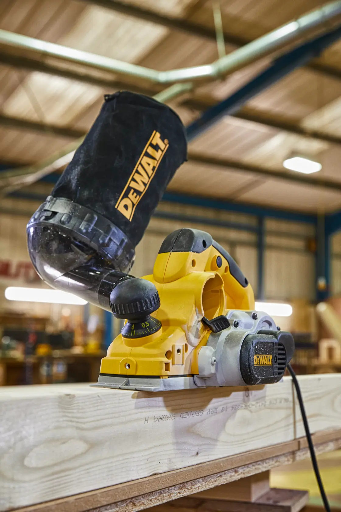 Nahaufnahme eines DEWALT Elektrohobels, der in einer Werkstatt auf einem Holzbrett verwendet wird. Der Hobel hat einen Staubauffangbeutel und der Hintergrund der Werkstatt ist unscharf.