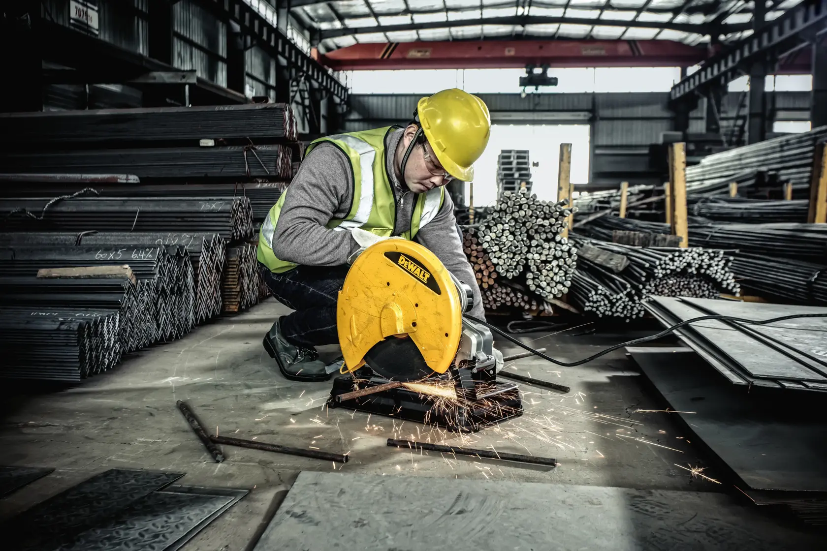 Un trabajador con casco de seguridad amarillo y chaleco reflectante está cortando metal con una sierra de corte amarilla DEWALT, produciendo chispas. El trabajador está en un almacén industrial rodeado de pilas de barras y láminas metálicas.