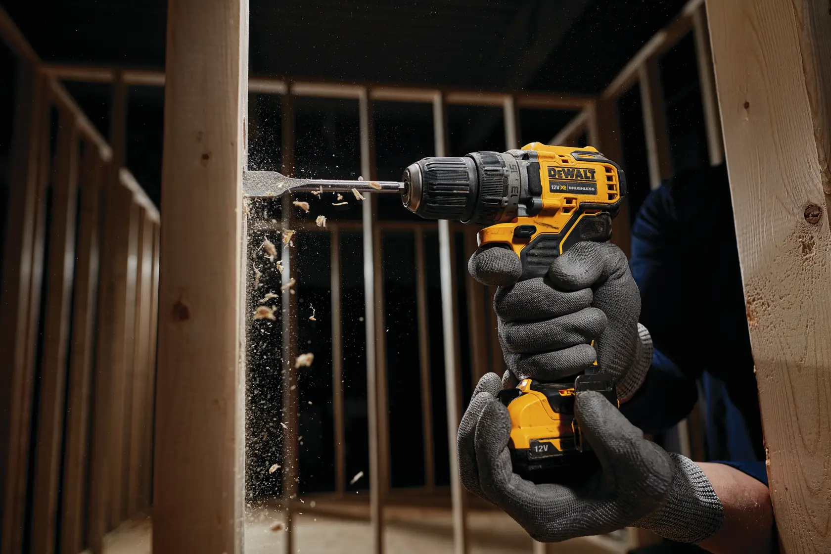 Eine Person mit Schutzhandschuhen benutzt einen gelben DEWALT Akkubohrer, um in einen Holzträger innerhalb einer Rahmenkonstruktion zu bohren. Holzspäne fliegen während des Bohrvorgangs.