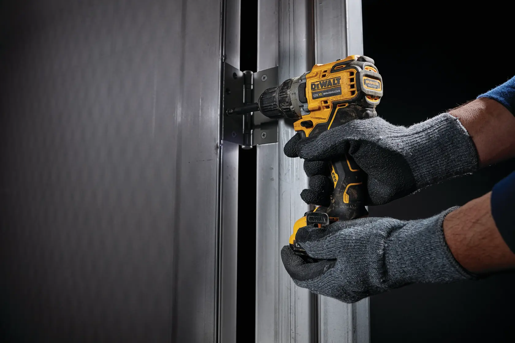 Eine Person mit grauen Handschuhen verwendet einen gelb-schwarzen DEWALT-Akkubohrer, um ein Scharnier an einem Metalltürrahmen zu befestigen.
