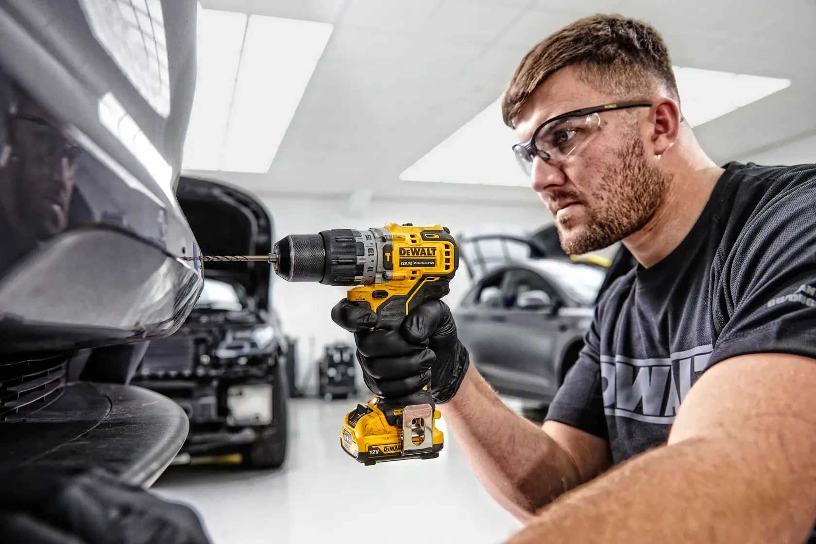 Una persona con guantes negros utiliza un taladro inalámbrico amarillo DEWALT DCD706D2 para perforar el parachoques de un coche en un taller automotriz. El rostro de la persona está desenfocado y hay varios coches visibles en el fondo.
