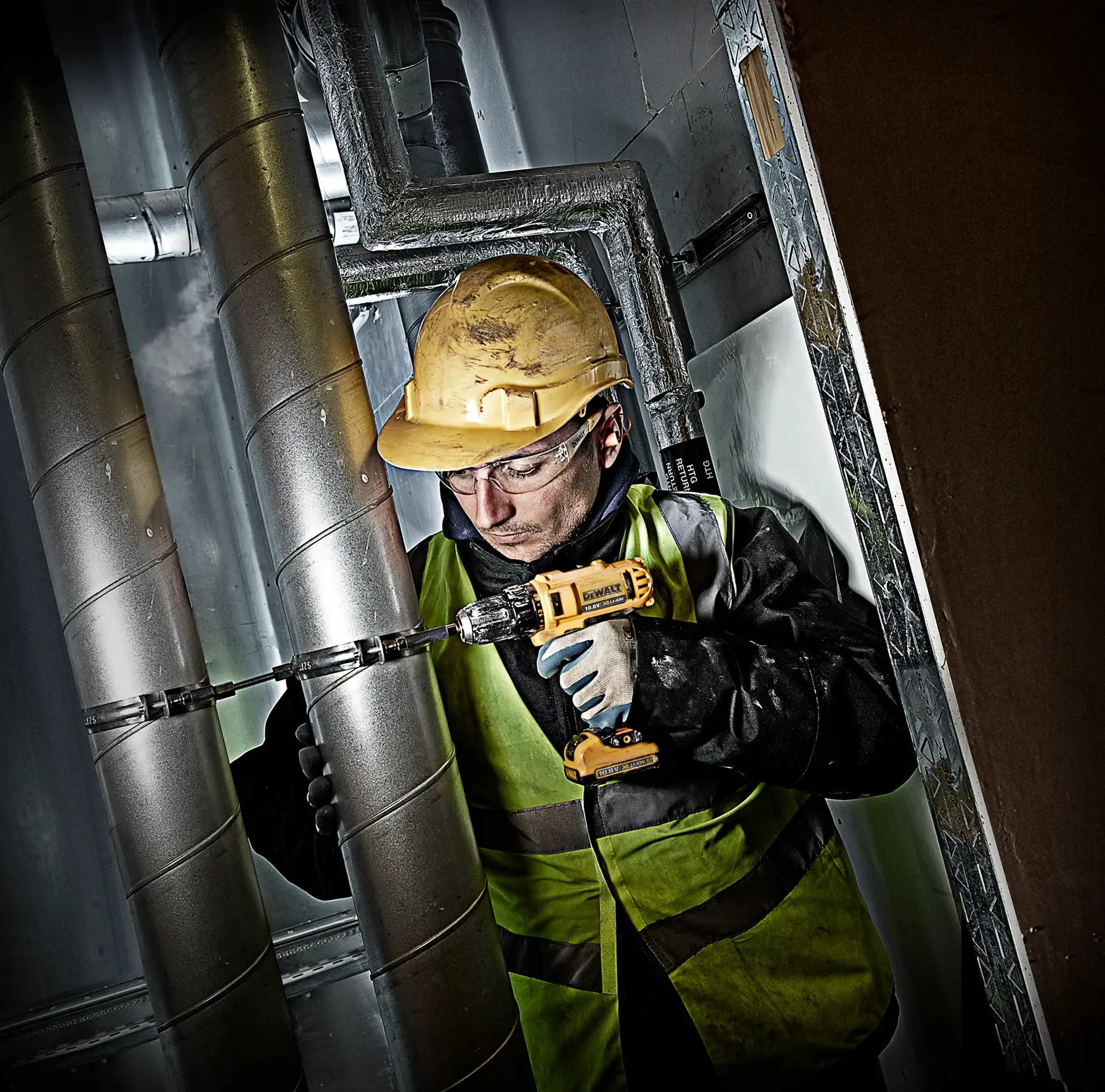 Worker in safety gear uses a DEWALT drill on metal pipes in an industrial setting.