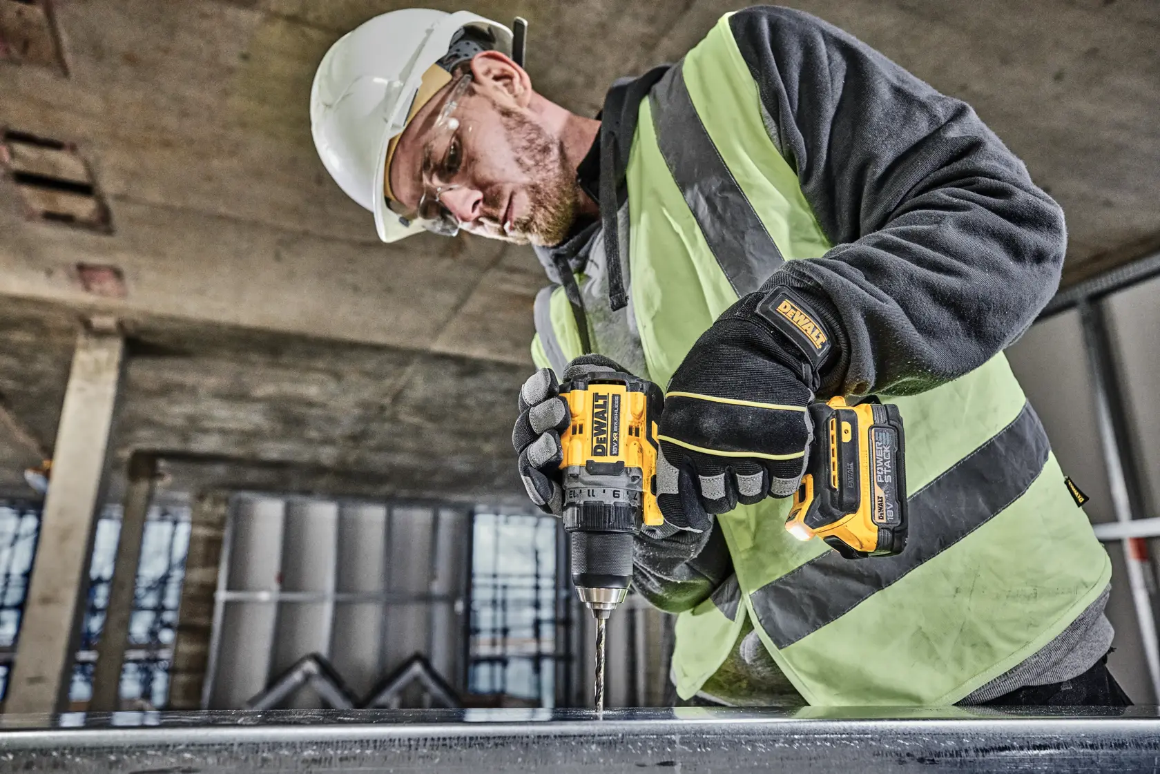 Eine Person mit Sicherheitsweste, Handschuhen und Helm benutzt einen DEWALT DCD800NT Akku-Bohrer, um eine Metalloberfläche auf einer Baustelle zu bohren.
