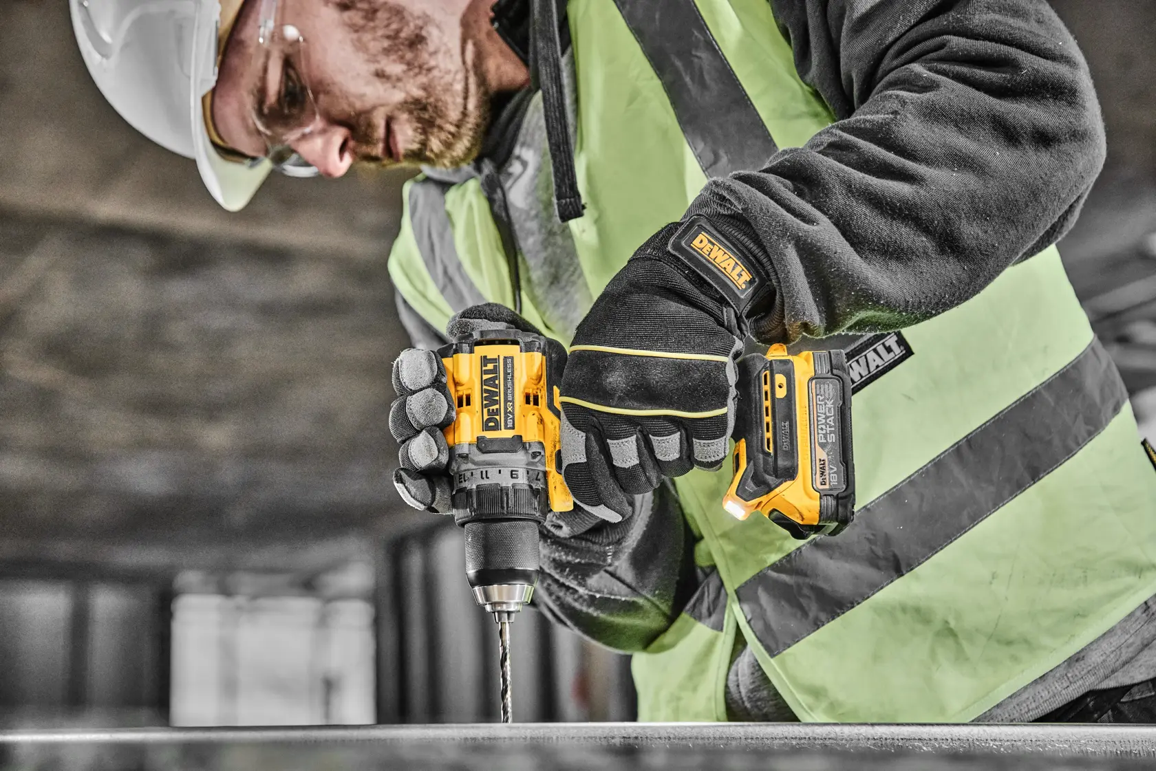 Eine Person mit Sicherheitsausrüstung, darunter Helm, Schutzbrille, Handschuhe und Warnweste, benutzt einen gelb-schwarzen kabellosen Akkubohrer von DEWALT auf einer Baustelle.