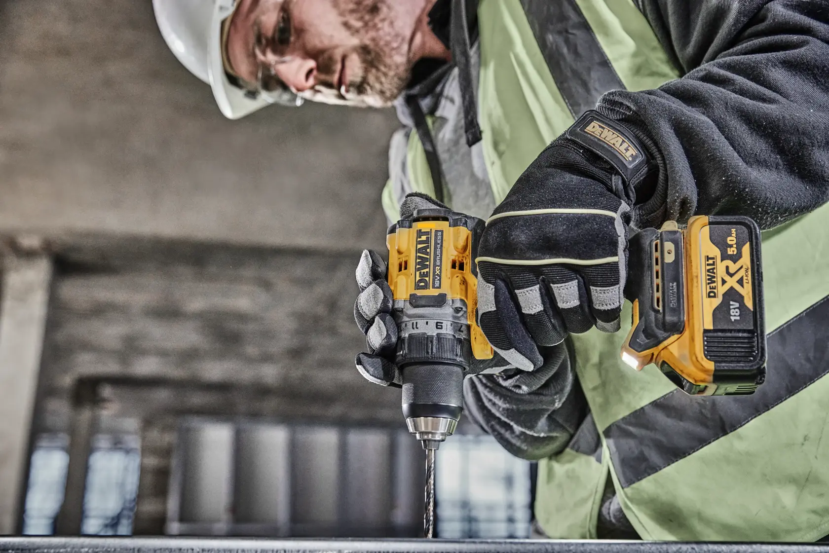 A person wearing safety gear operates a DEWALT DCD800P2T cordless drill with an 18V XR battery, drilling into a metal surface at a construction site.