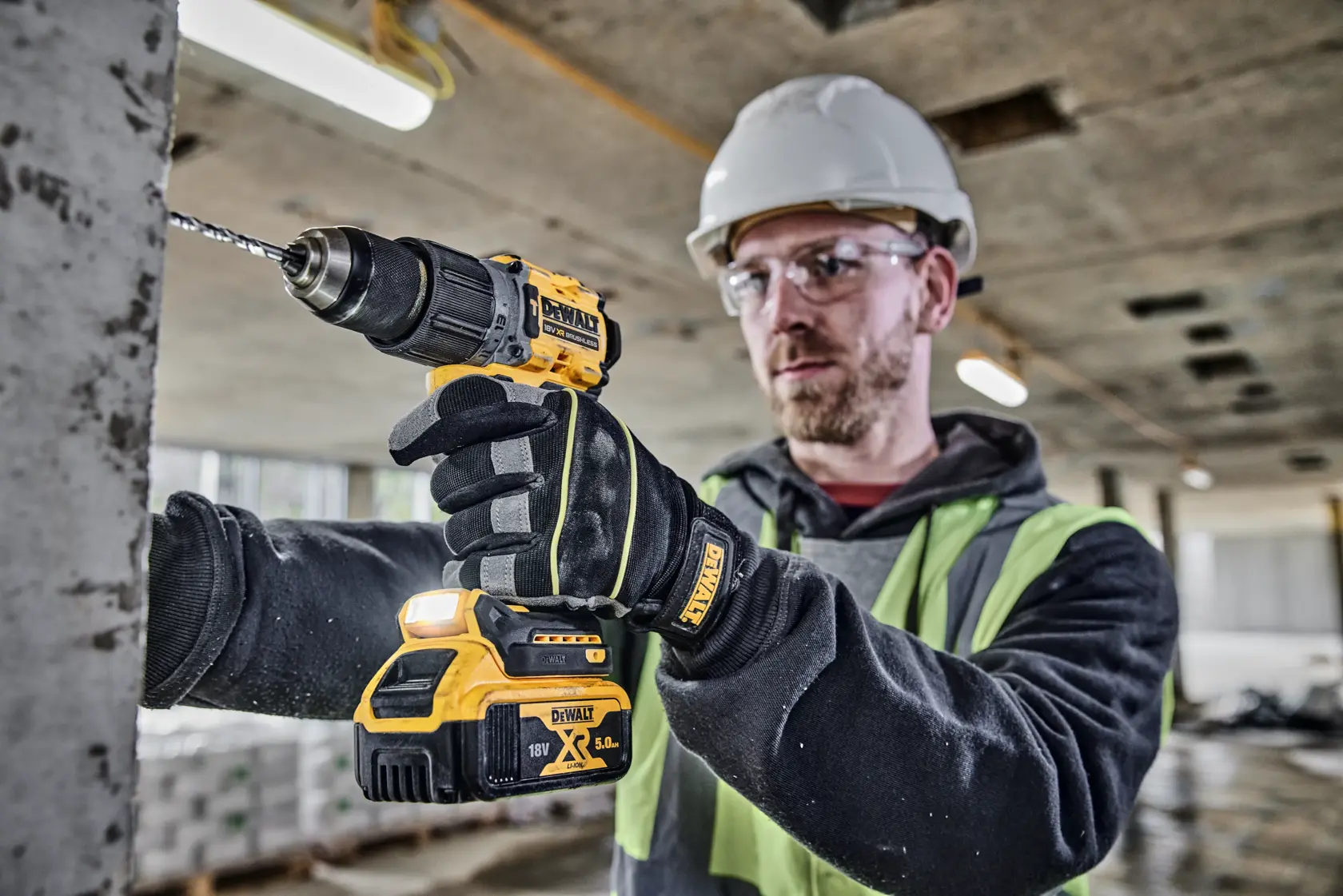 Una persona con guanti, giubbotto di sicurezza e casco sta usando un trapano a batteria DEWALT DCD805P2T per forare una superficie di cemento in un cantiere. Il volto è sfocato per privacy.