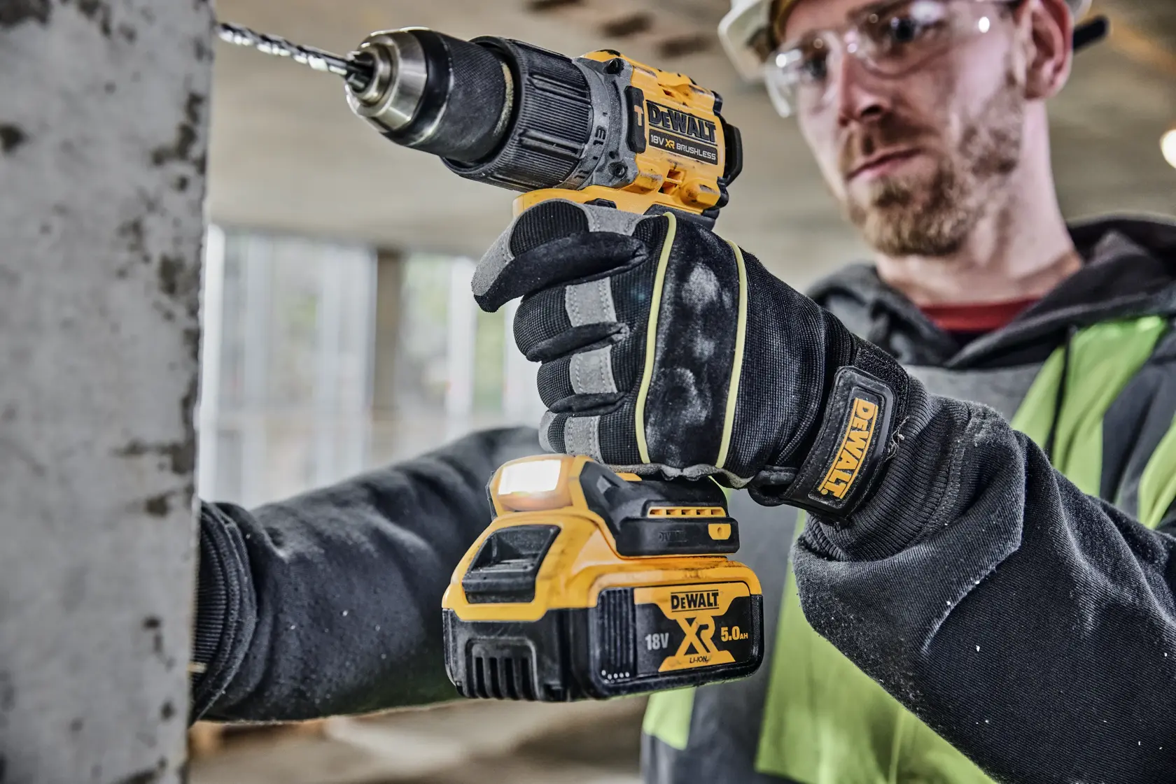 Una persona che indossa guanti DEWALT sta utilizzando un trapano avvitatore a batteria DEWALT 18V XR (modello DCD805P2T) per forare una superficie di cemento in un ambiente di cantiere interno. Il volto della persona è sfocato.