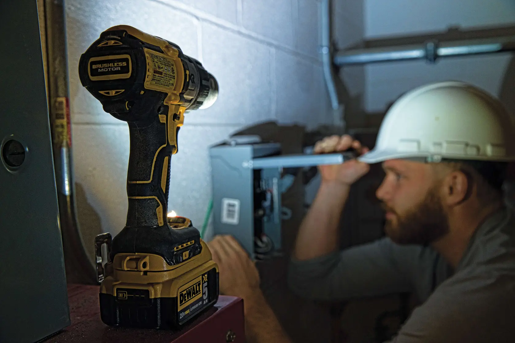 A person wearing a high-visibility jacket and hard hat is using a DEWALT cordless power drill (model DCD996P2) to drill into a concrete or masonry wall in an industrial or construction setting. The product and grip on the tool are clearly visible.