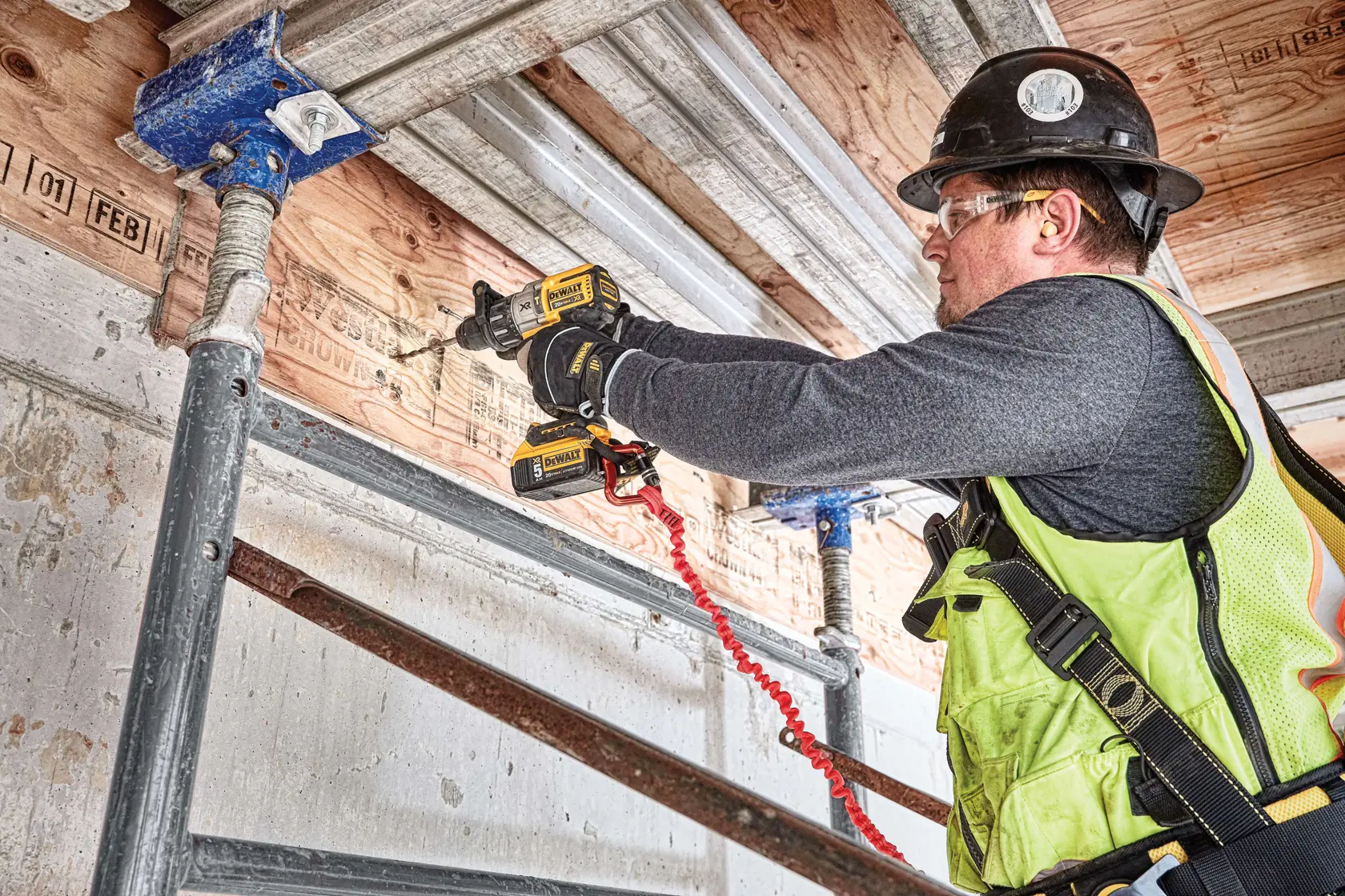 A person using a DEWALT cordless drill (model DCD996P2) to drill into a wooden beam at a construction site.