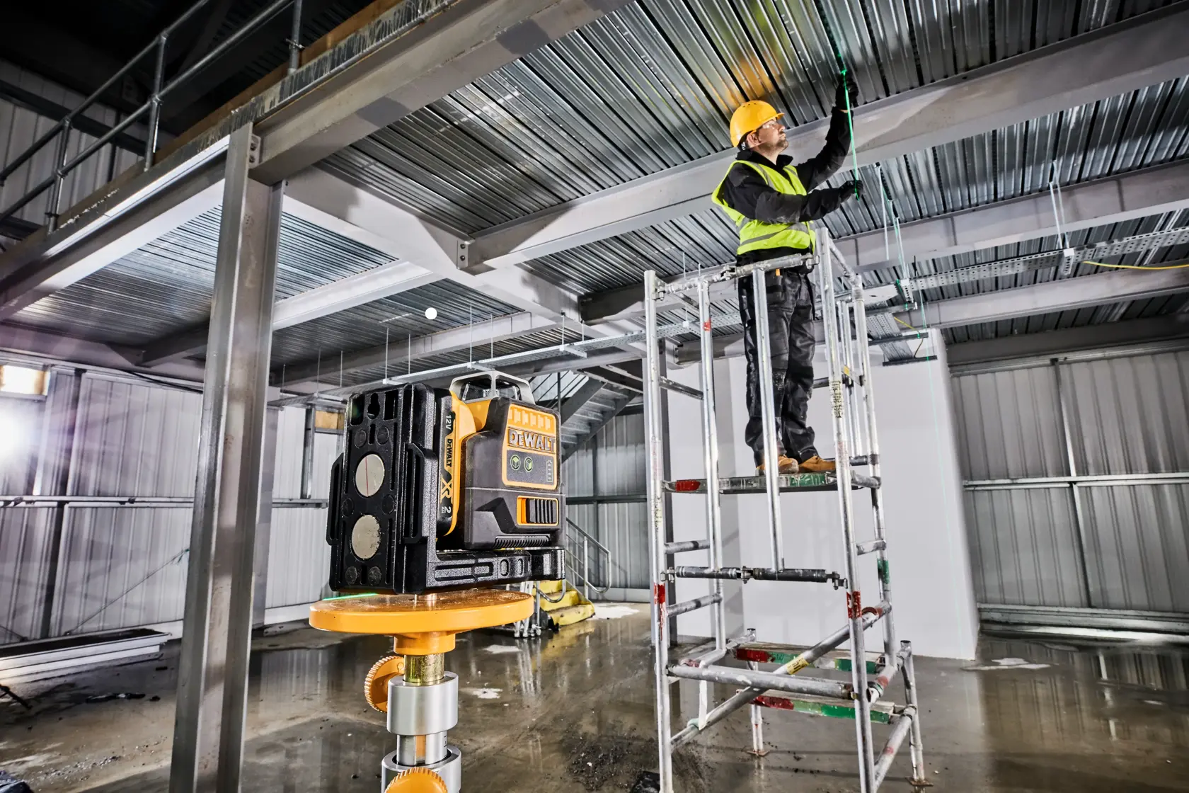 Ein DEWALT-Laser-Nivelliergerät ist auf einem Stativ in einem Industriegebäude im Bau eingerichtet. Im Hintergrund steht ein Arbeiter mit Sicherheitsweste und Helm auf einem Gerüst und markiert oder arbeitet an der Metalldecke.