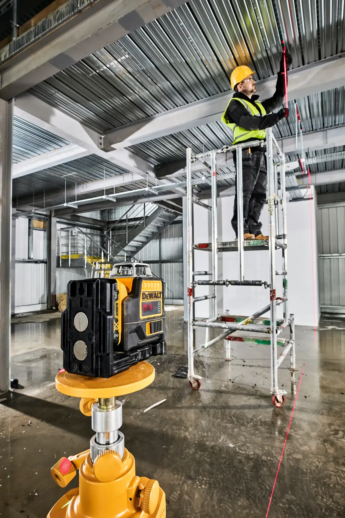 Ein DEWALT-Lasernivelliergerät steht auf einem Stativ im Vordergrund einer industriellen Baustelle. Im Hintergrund steht ein Arbeiter mit gelbem Schutzhelm und Warnweste auf einem Gerüst und markiert eine Metalldecke entlang der sichtbaren roten Laserlinie des Geräts. Der Bereich zeigt Metallträger, Treppen und verstreute Baumaterialien.