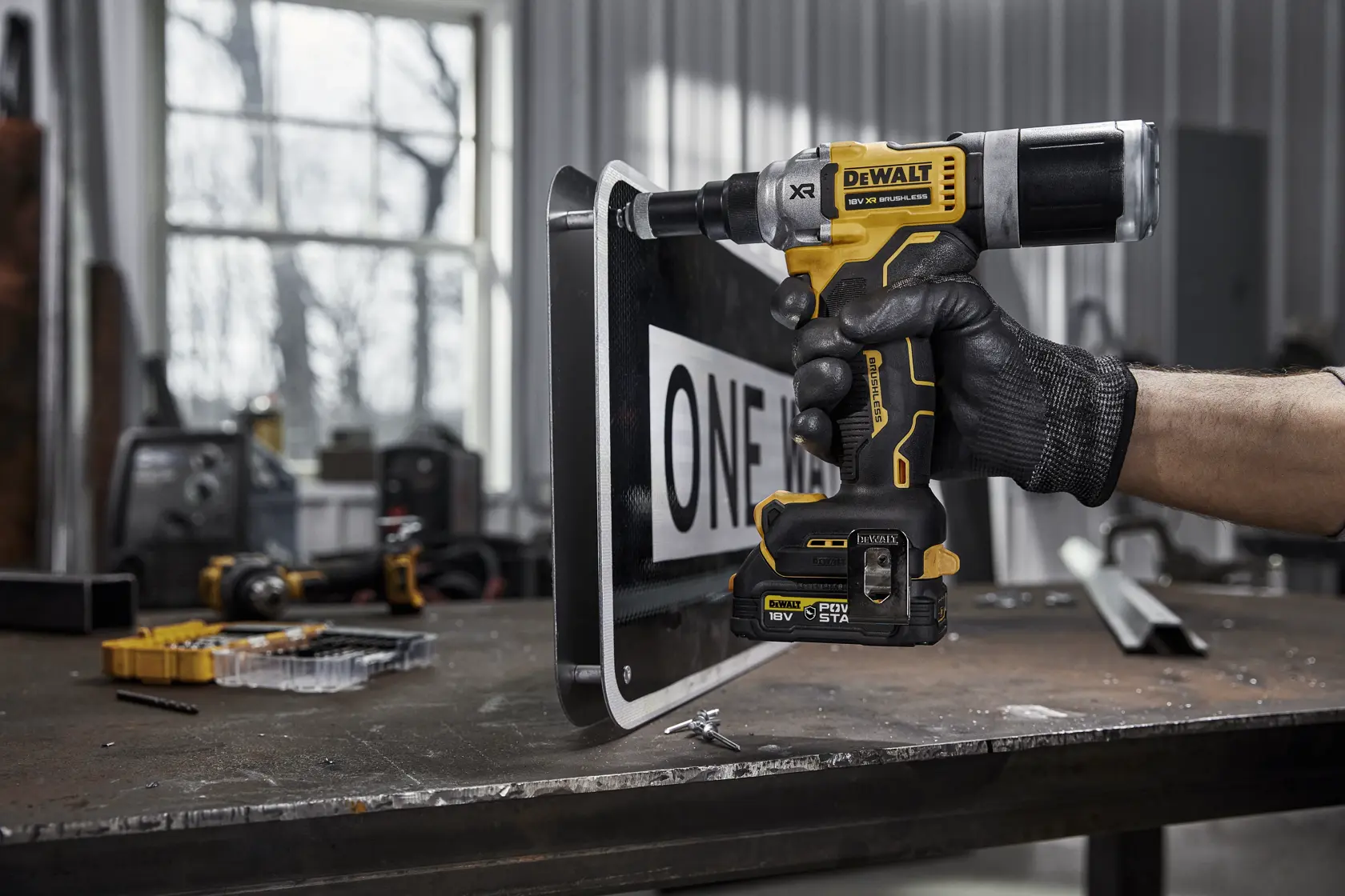 Una persona con un guante negro utiliza una herramienta eléctrica inalámbrica DEWALT DCF414NT sobre una señal metálica en un taller. La herramienta muestra el logotipo amarillo y negro de DEWALT y está colocada contra el borde de la señal. Se pueden ver varias herramientas y piezas en el banco de trabajo al fondo.