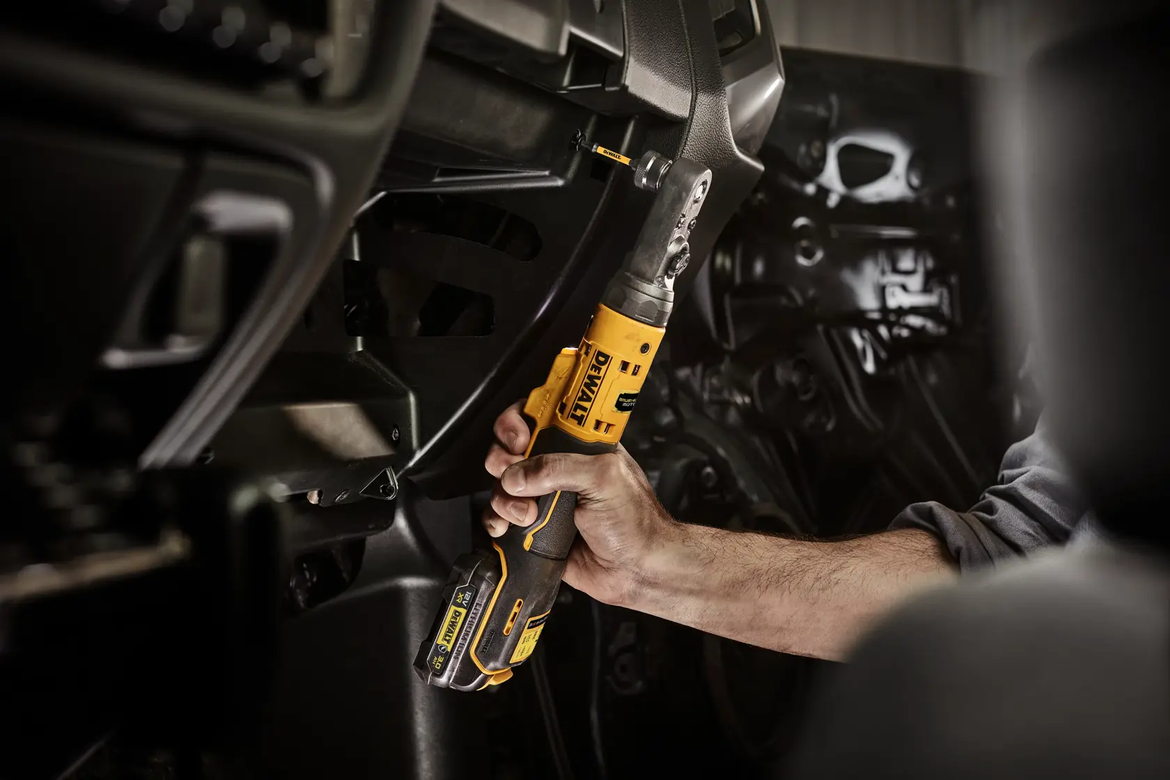 Mano usa herramienta de carraca inalámbrica DEWALT amarilla dentro de un vehículo para trabajo mecánico.