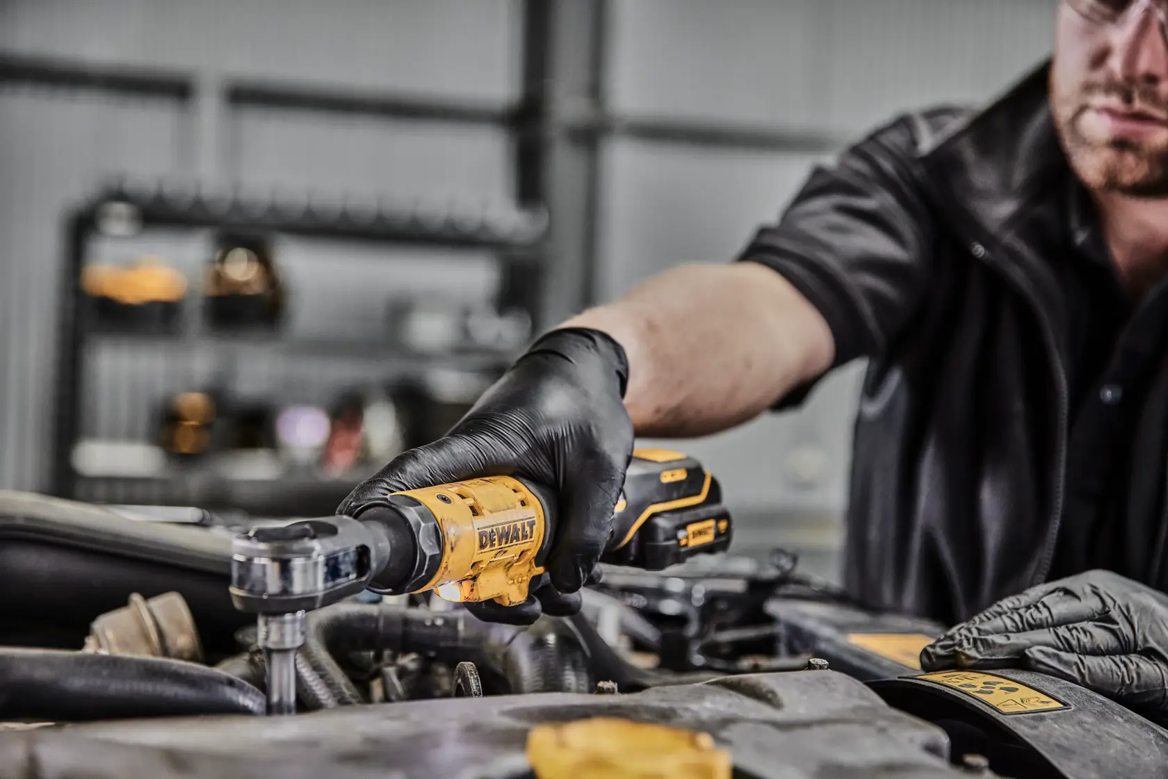 Eine Person mit schwarzen Handschuhen verwendet ein gelbes kabelloses Ratschenwerkzeug von DEWALT, um an einem Motor in einer Werkstatt zu arbeiten.