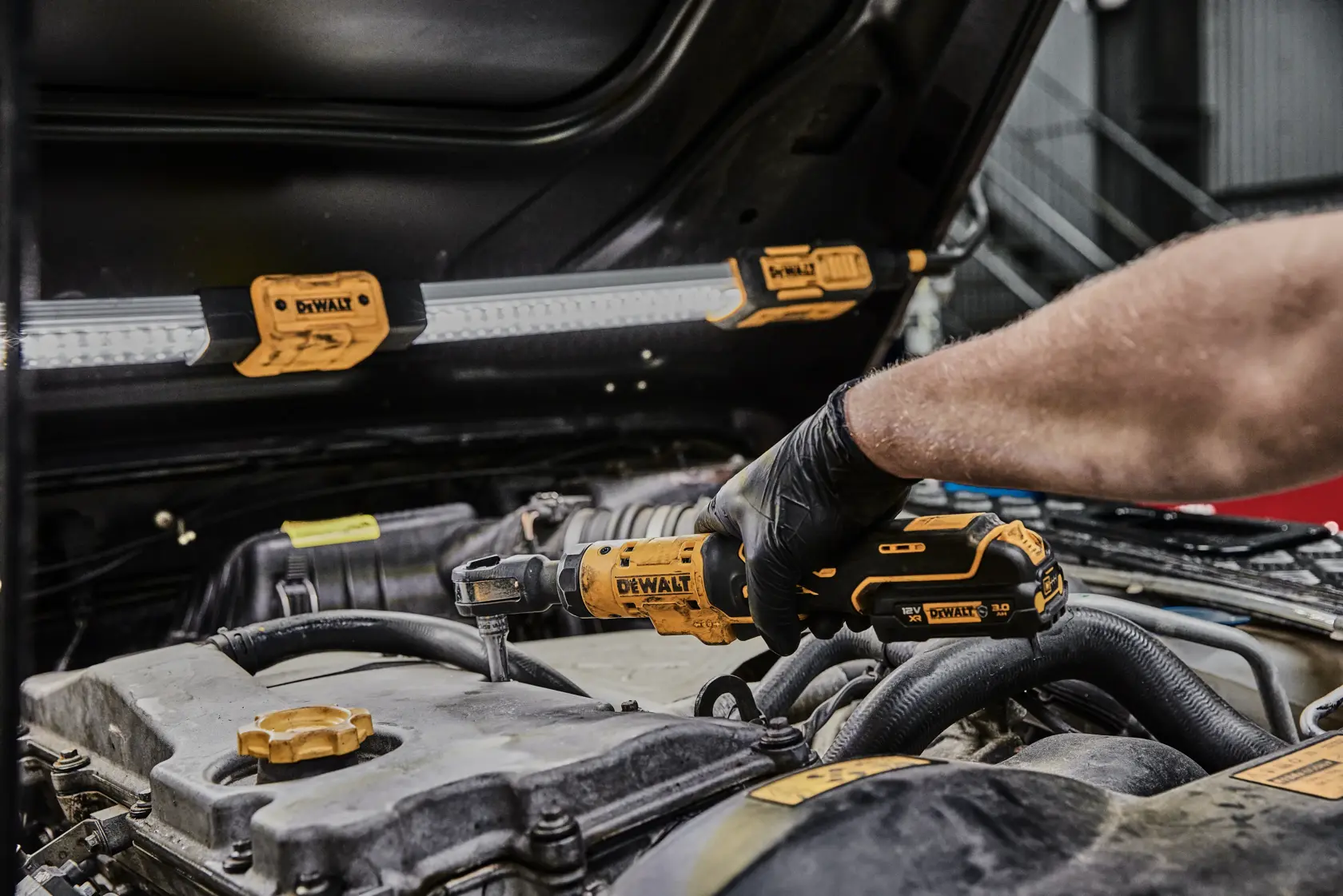 Una persona utiliza una llave de carraca inalámbrica DEWALT (modelo DCF504N) para trabajar en el motor de un coche en un taller. Una luz de trabajo LED con la marca DEWALT está sujeta al capó para iluminación.