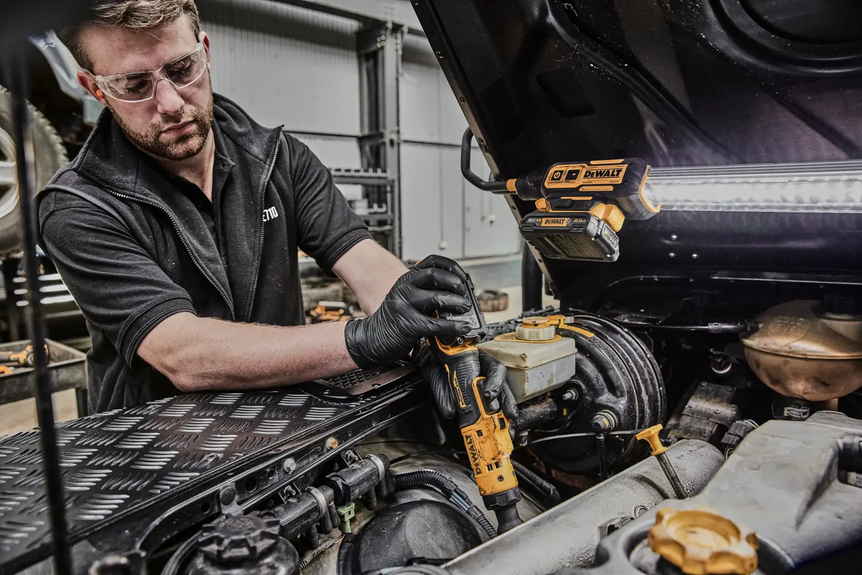 Una persona con guantes negros utiliza una herramienta eléctrica DEWALT, probablemente una carraca inalámbrica, para trabajar en el motor de un vehículo en un taller. Una lámpara de trabajo DEWALT está fijada debajo del capó abierto del coche, iluminando el área de trabajo.