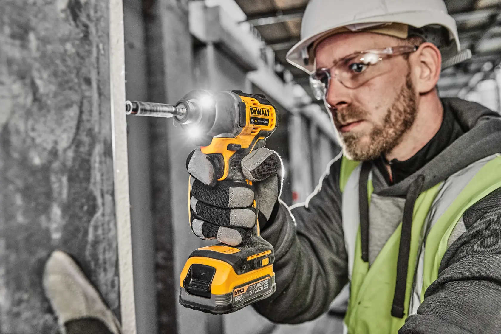 A person wearing safety gear uses a DEWALT DCF840N cordless impact driver to fasten a screw to a wall in a construction setting. The person’s face is blurred for privacy.