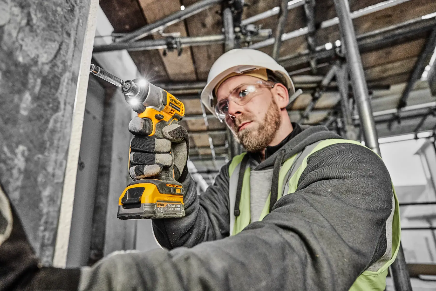 A person wearing work gloves, a hard hat, and a high-visibility vest is using a DEWALT DCF840N cordless impact driver to drive a screw into a metal surface on a construction site. The tool is yellow and black, and the work environment contains scaffolding and metal structures.