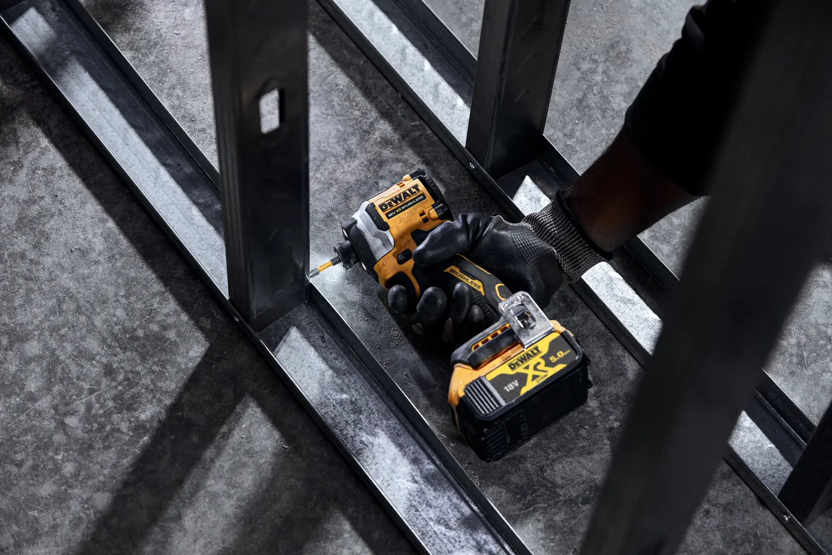 Mano enguantada usa atornillador de impacto DEWALT inalámbrico en marco de acero.