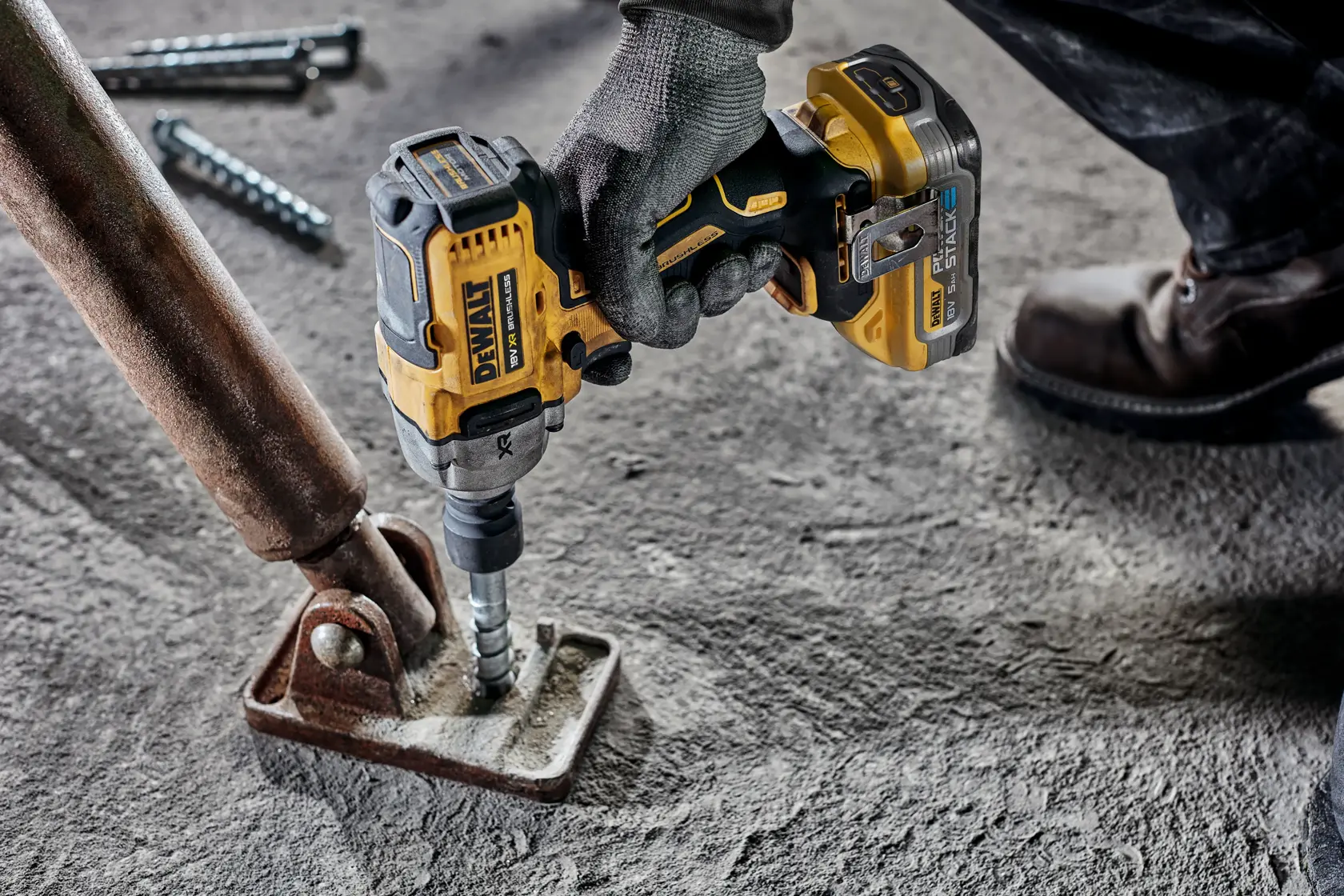 Una persona con guantes de trabajo y botas utiliza una llave de impacto inalámbrica DEWALT para apretar un perno en un soporte de metal oxidado fijado a una superficie de hormigón.