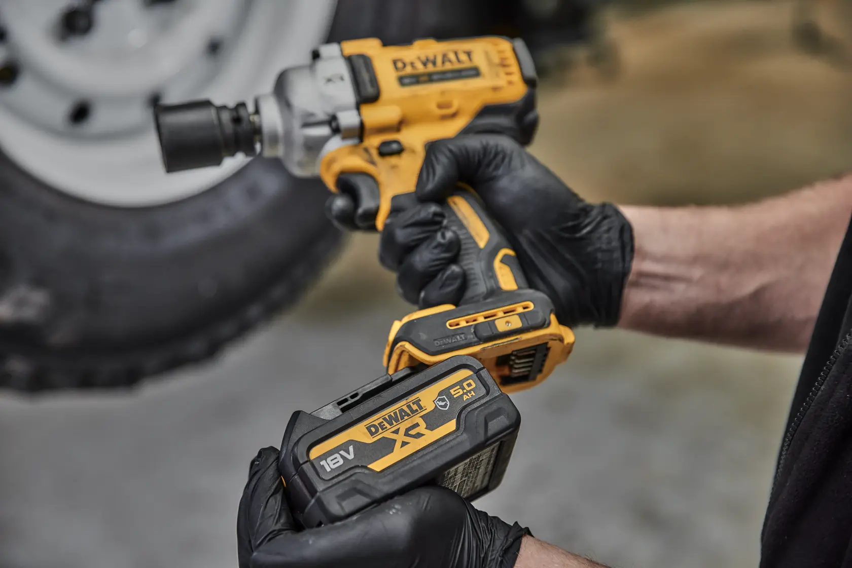Person wearing gloves holds a DEWALT cordless impact wrench and 18V battery near a vehicle wheel.