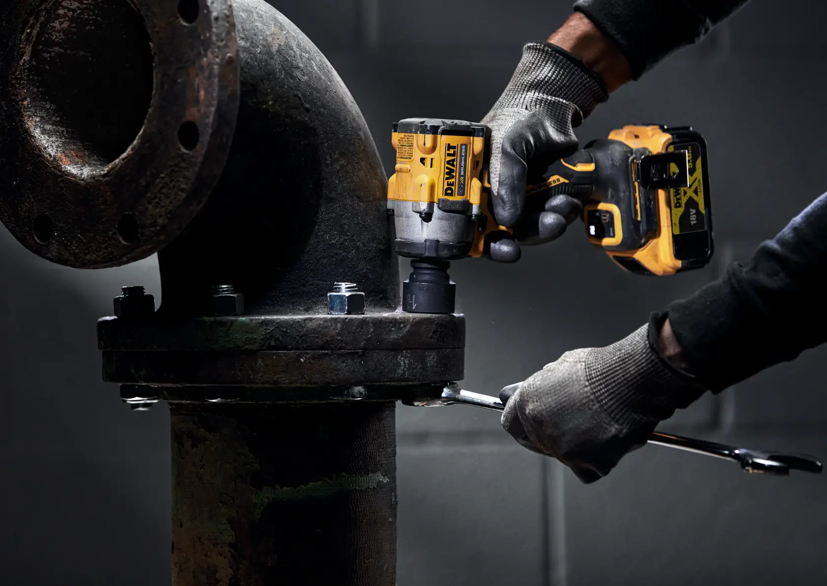 A person wearing work gloves uses a DEWALT DCF922P2T cordless impact wrench to tighten bolts on a large industrial pipe joint.