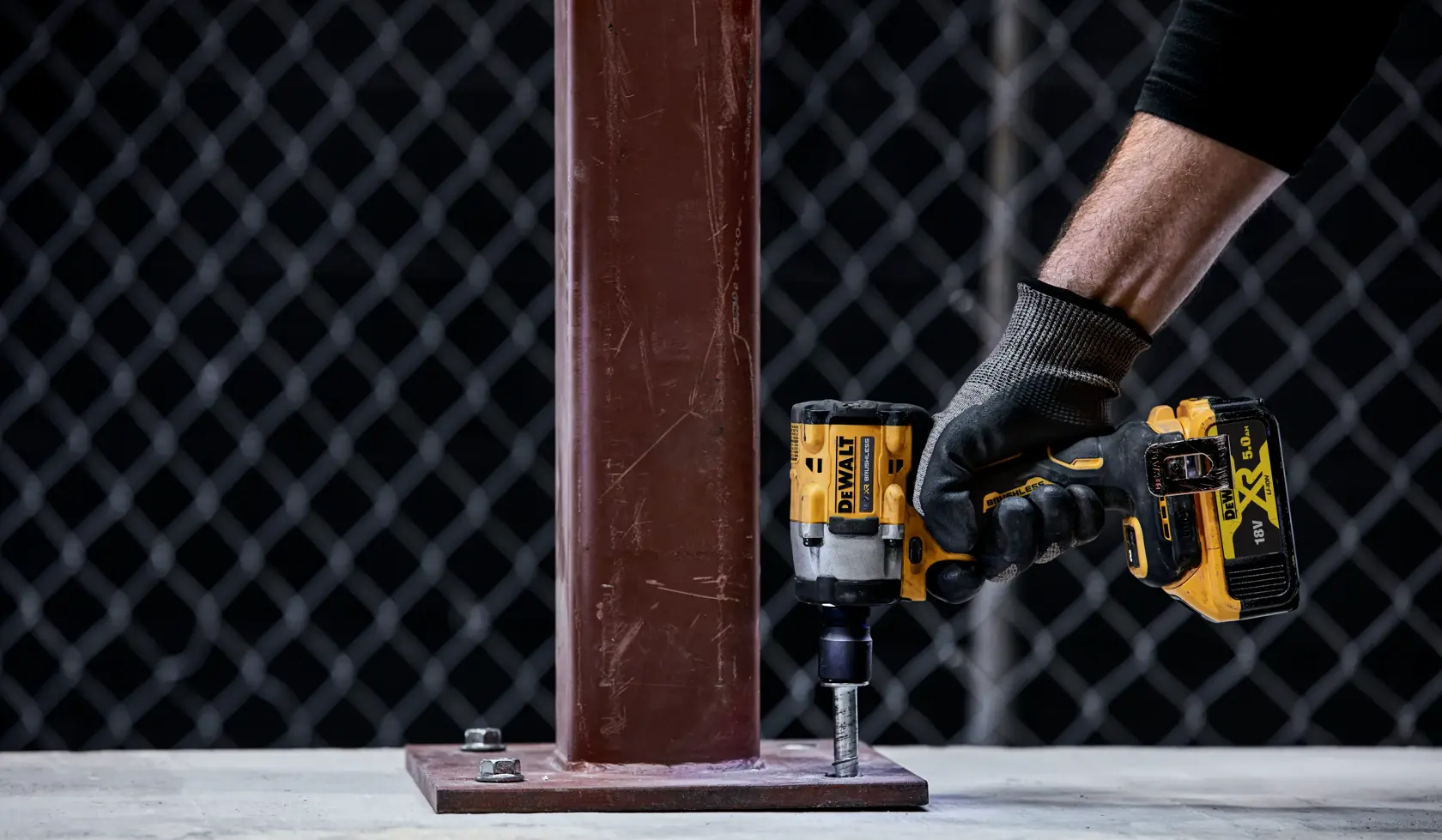A person wearing a glove is using a DEWALT cordless impact wrench to tighten a bolt on a metal base plate attached to a vertical steel beam.