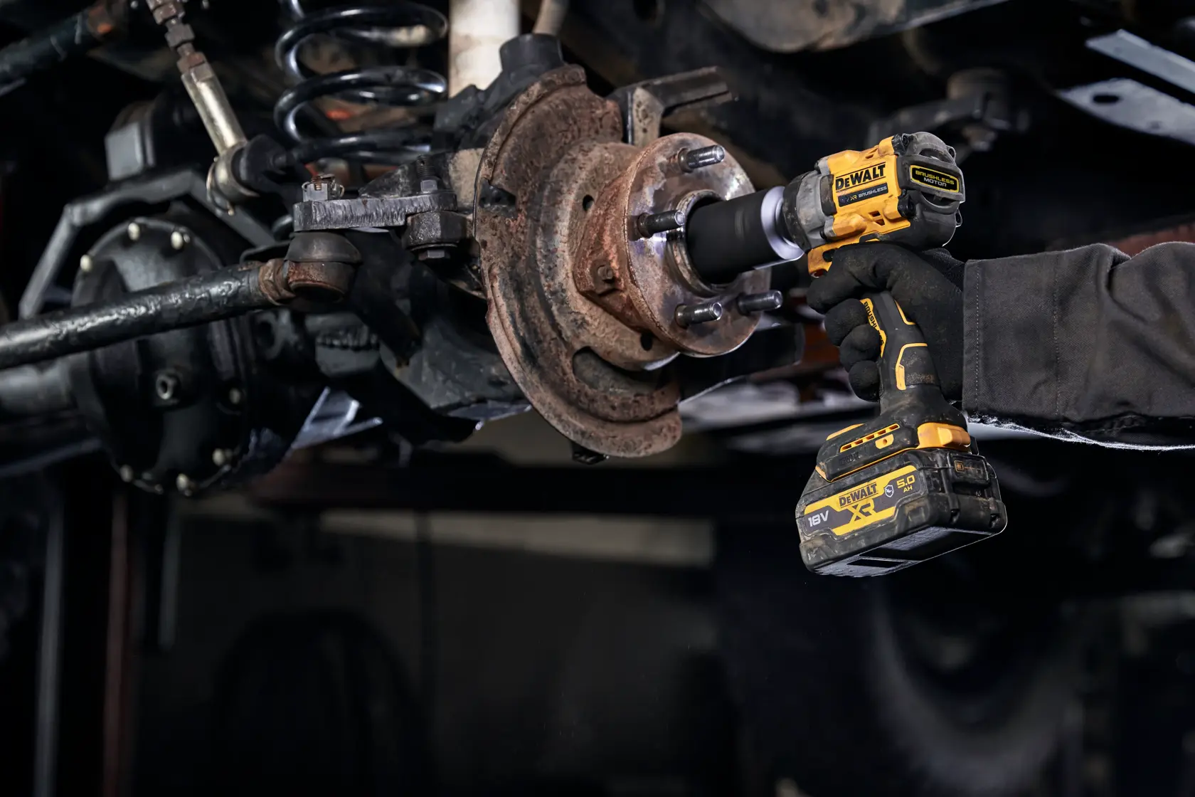 Ein DEWALT DCF923P2G Akku-Schlagschrauber wird verwendet, um an einer rostigen Radnabe unter einem Fahrzeug zu arbeiten. Das Werkzeug wird von einer behandschuhten Hand gehalten.