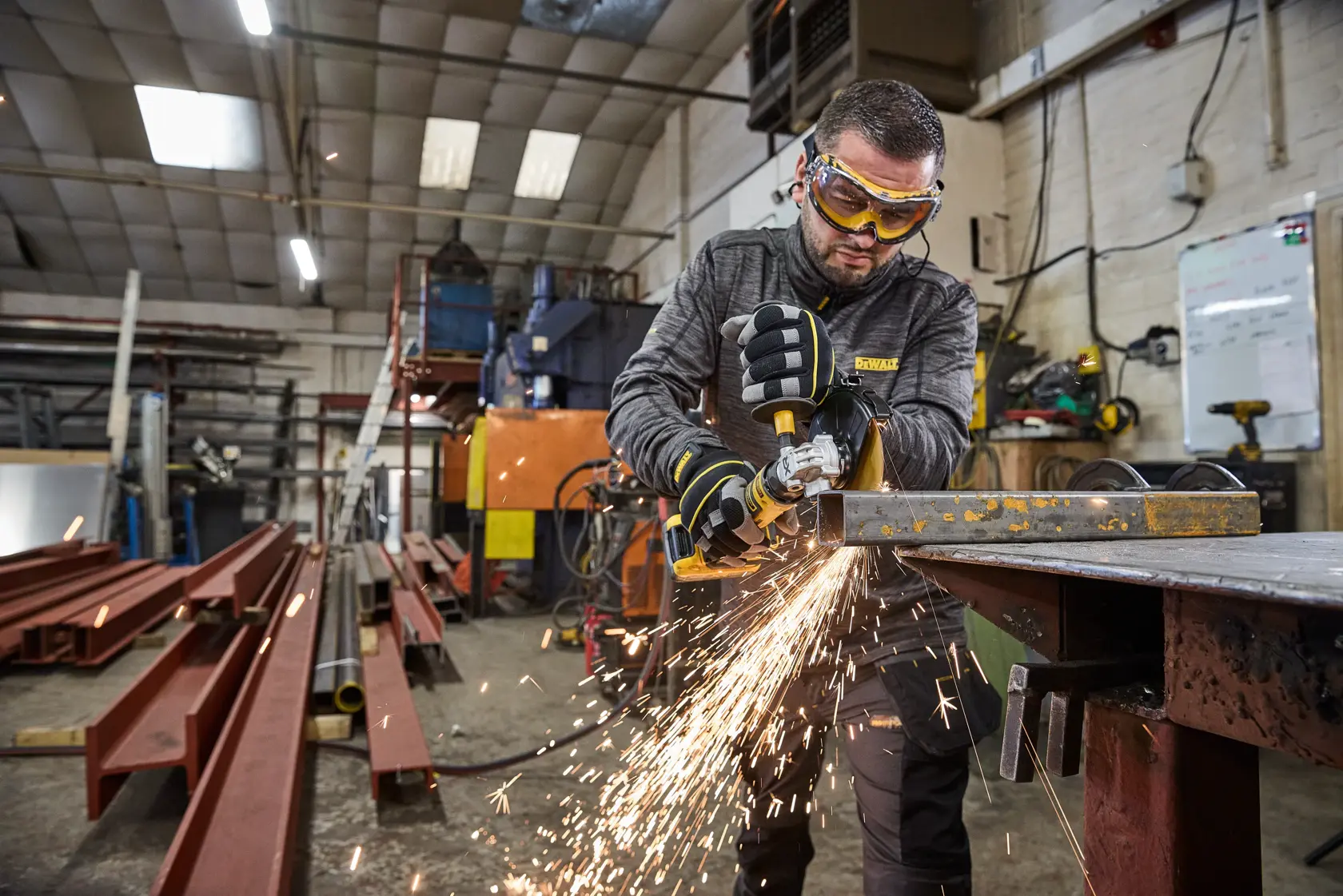 Persona usando una amoladora DEWALT en metal en taller, salen chispas. Hay equipamiento industrial alrededor.