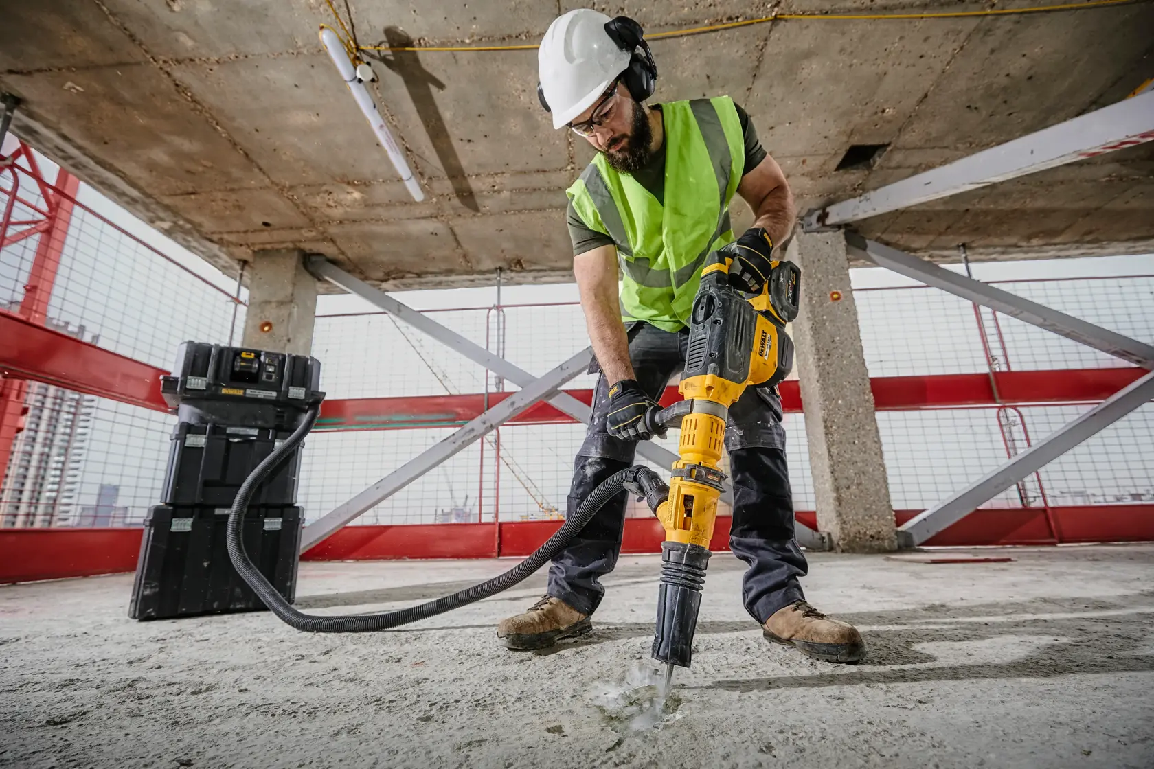 Un operaio edile con gilet di sicurezza, casco e guanti utilizza un martello perforatore DEWALT DCH773Y2 per rompere il cemento in un cantiere. Il trapano è collegato a un sistema di aspirazione polvere e una valigetta DEWALT è visibile sullo sfondo.