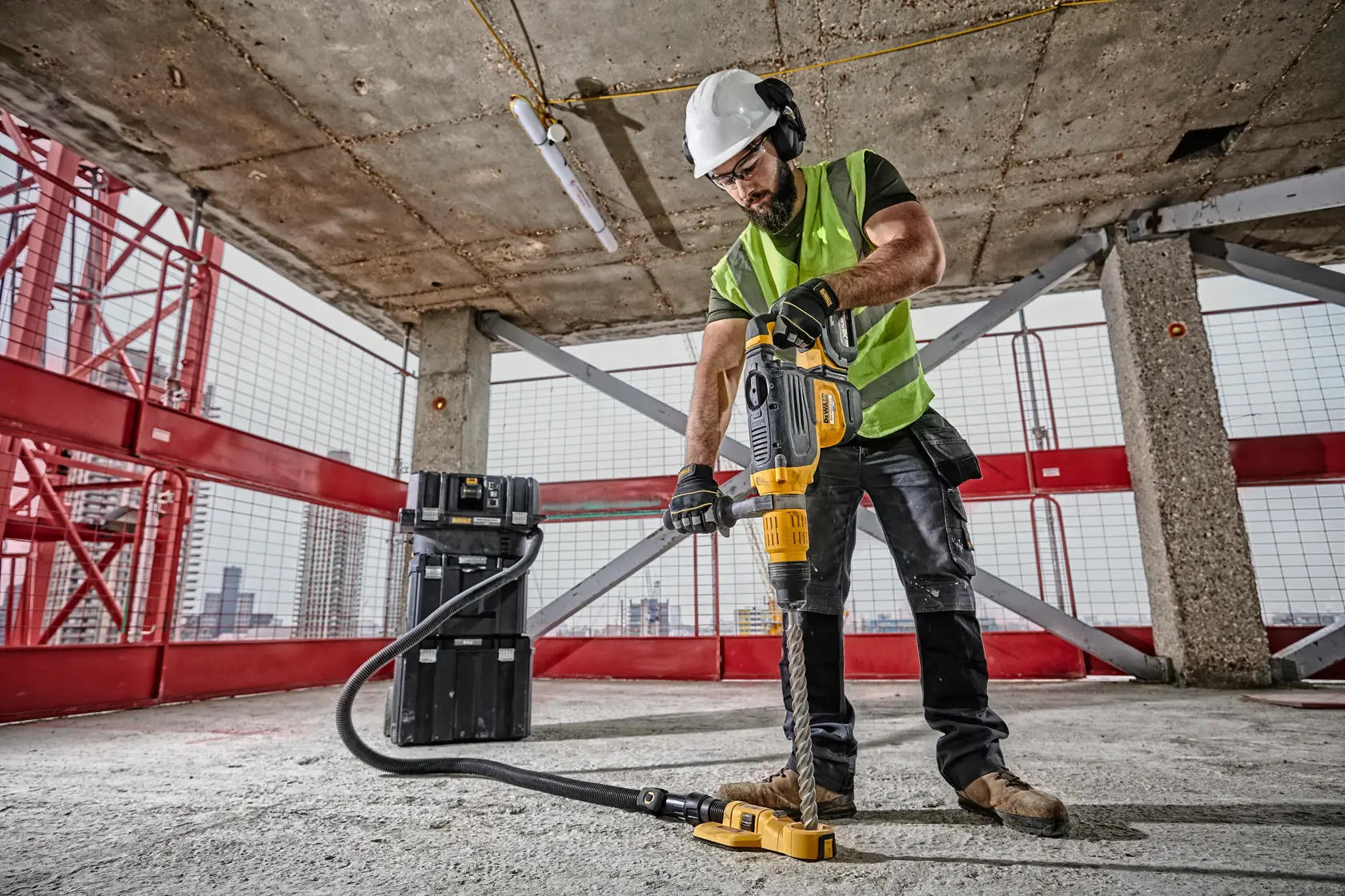 Un operaio edile con casco di sicurezza e gilet ad alta visibilità utilizza un martello rotante giallo DEWALT su un pavimento di cemento in un cantiere, con un aspiratore collegato e una valigetta nera vicina.
