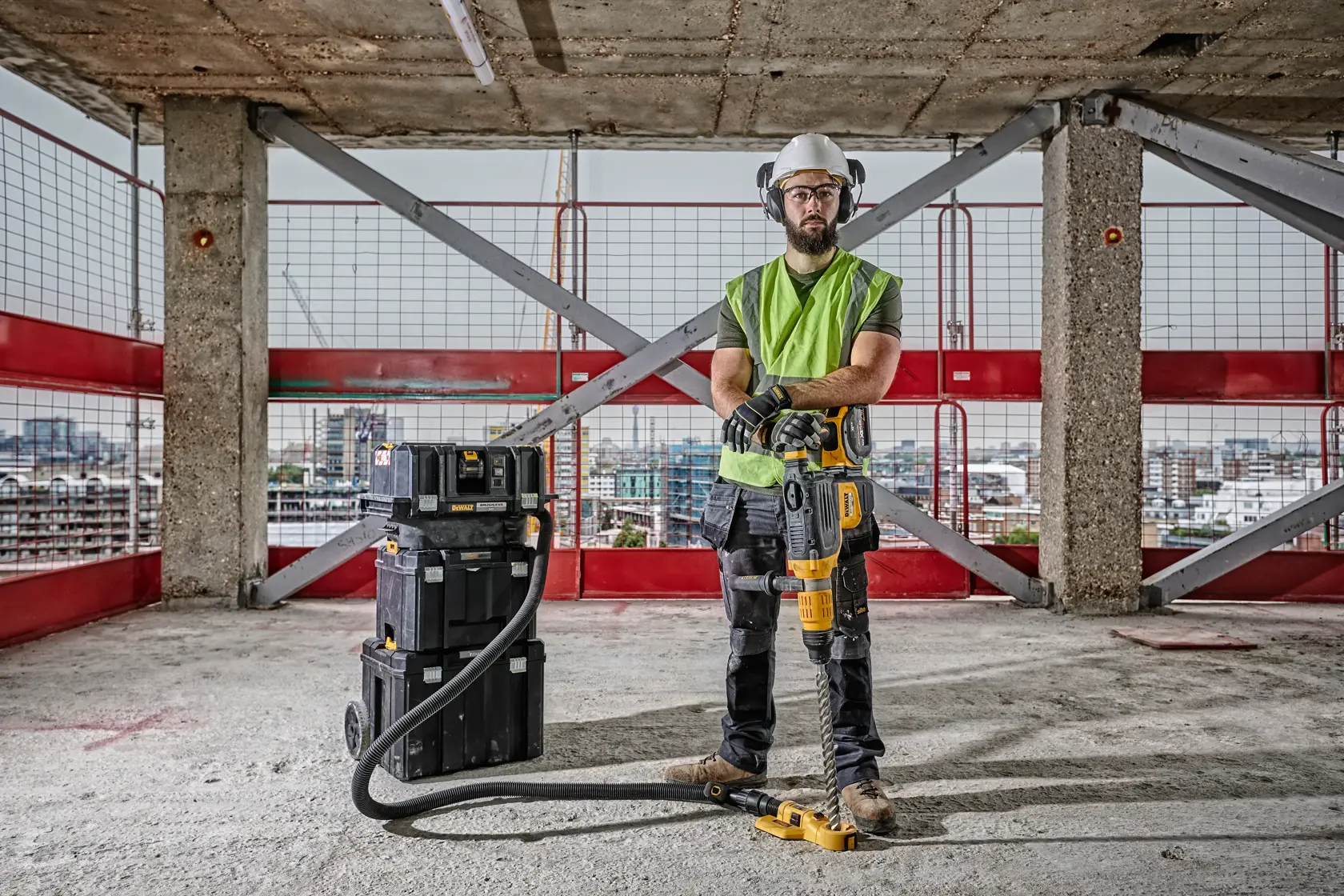 Un operaio edile con equipaggiamento di sicurezza utilizza un martello rotante DEWALT DCH773Y2 collegato a una scatola porta attrezzi tramite un tubo, su un pavimento di cemento all'interno di un edificio non finito.