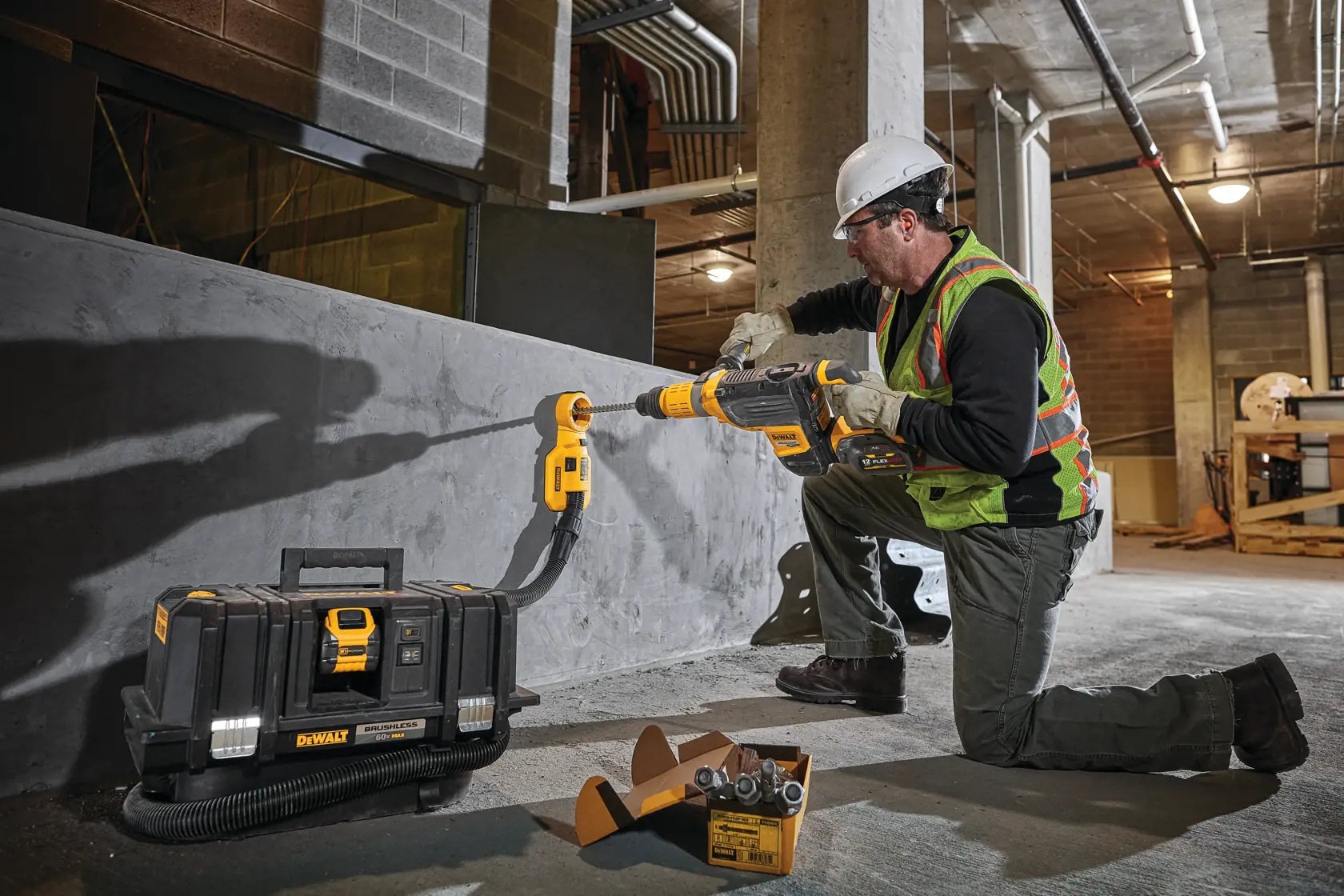 Un operaio indossa dispositivi di sicurezza, tra cui un casco e un gilet ad alta visibilità, e utilizza un martello perforatore DEWALT DCH773Y2 per forare un muro di cemento in un cantiere. Un estrattore di polvere DEWALT e una valigetta degli attrezzi si trovano nelle vicinanze, insieme a una scatola di punte da trapano.