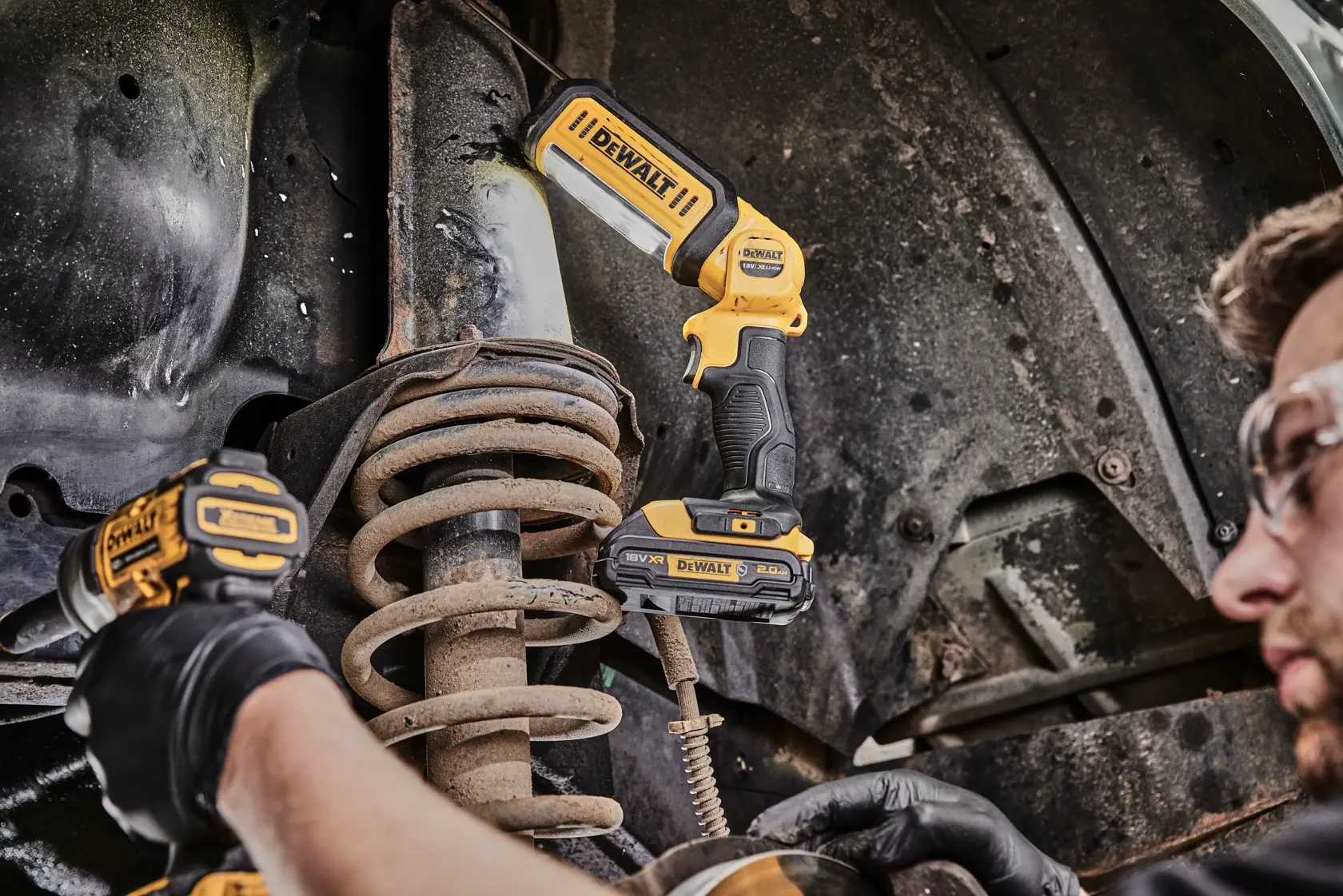 A person is kneeling and working on an open electrical unit, using tools. On the floor is a DEWALT DCL050 portable LED work light, illuminating the workspace.