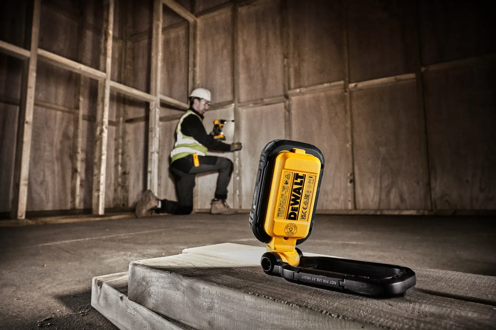 DEWALT DCL182 portable work light placed on a construction site floor, with a person in safety gear kneeling in the